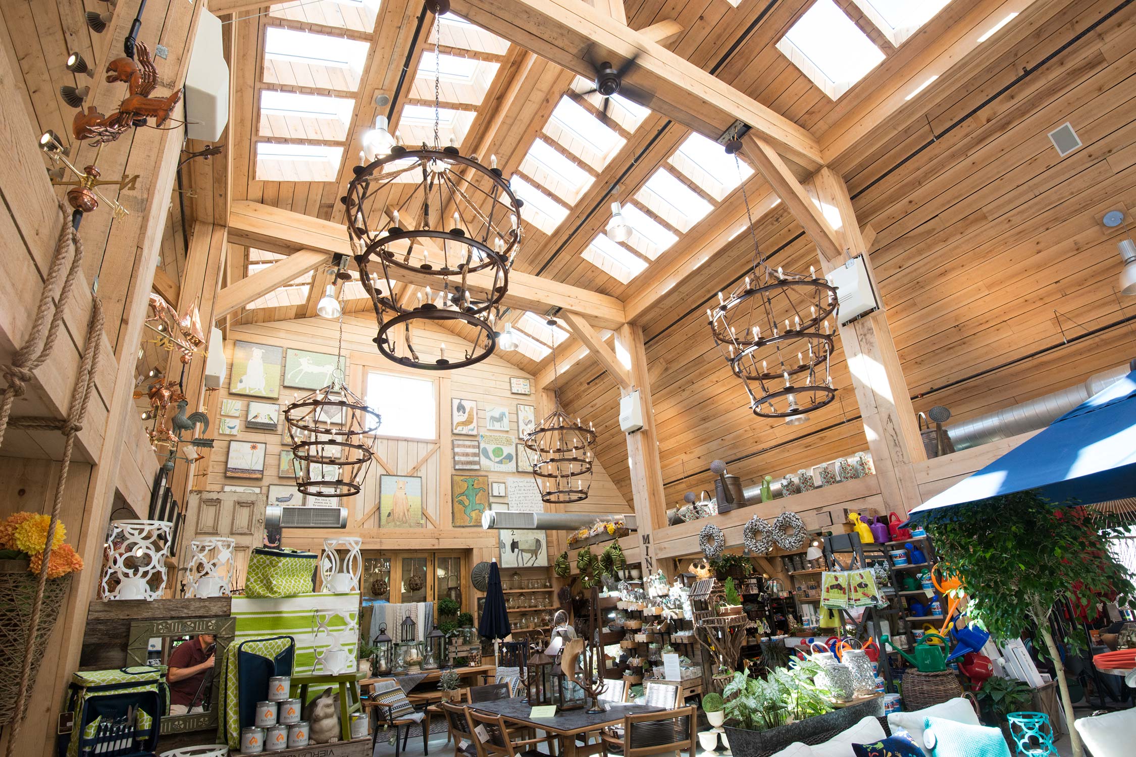 Rustic barn interior with wooden beams, chandeliers, skylights, and decor items like plants, pillows, and garden supplies.
