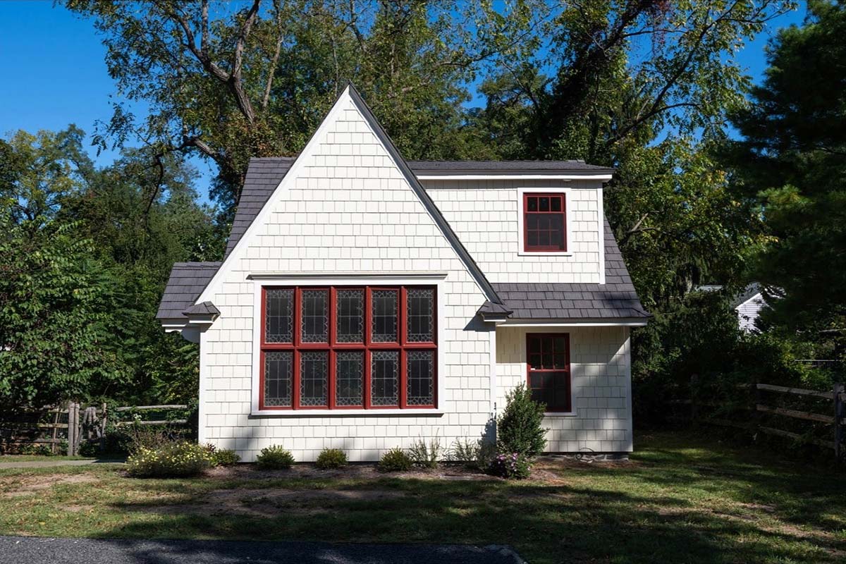A white house with a steep roof, large decorative windows with red frames, and a small garden, surrounded by trees.