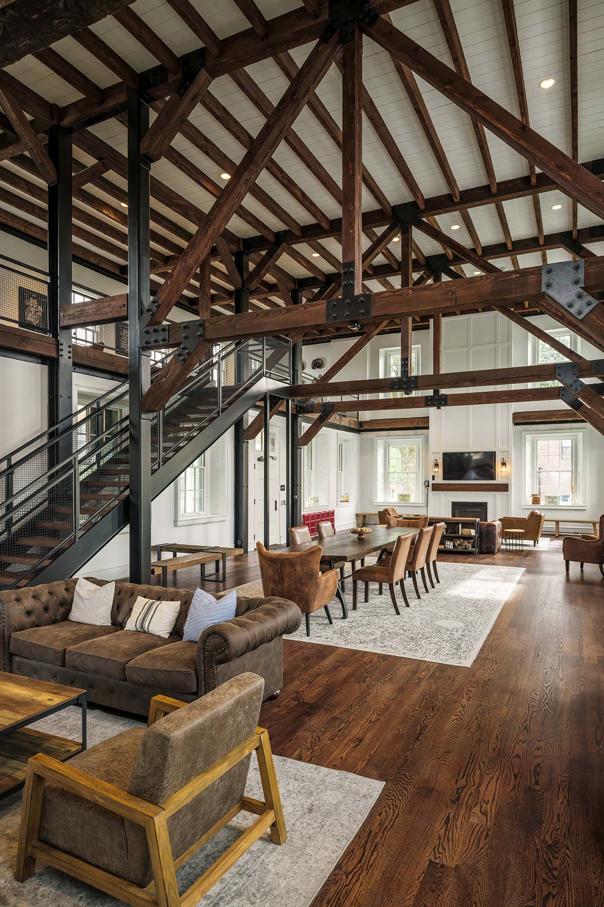 Large room with wooden beams, metal staircase, dining table, leather chairs, sofa, and fireplace with TV.