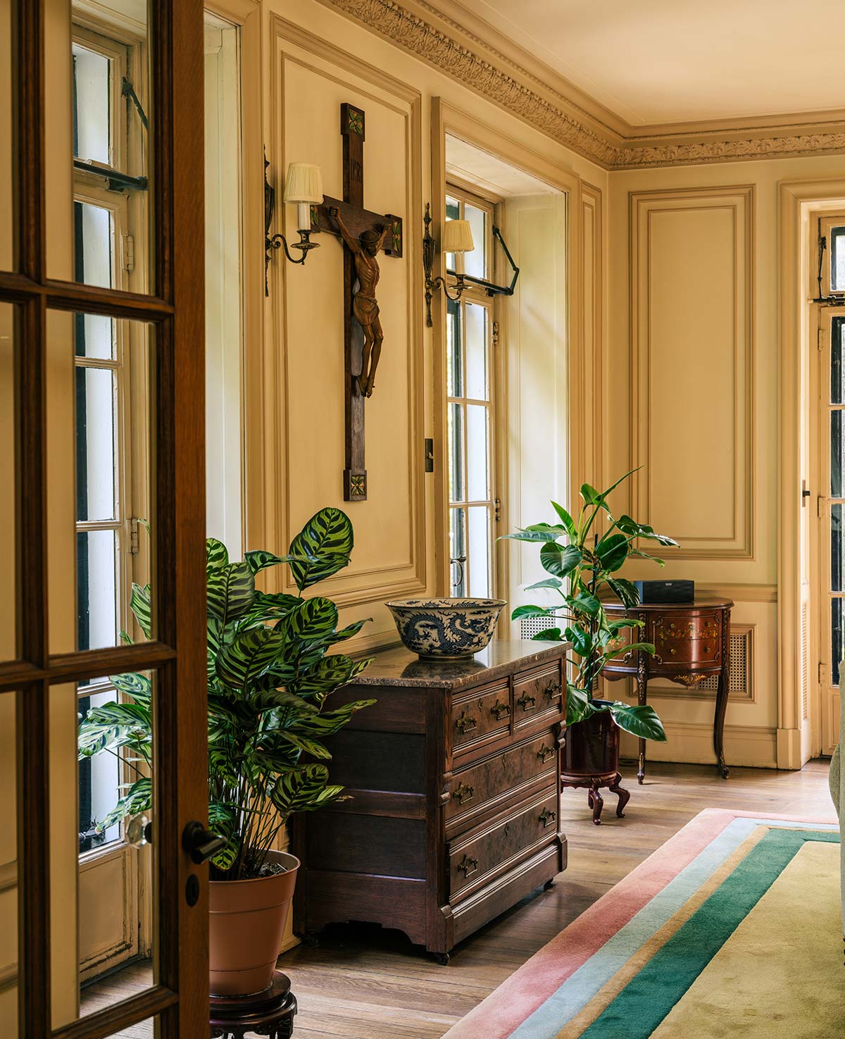 Elegant room with wood-paneled walls, a crucifix, and potted plants. Features a wooden dresser with a decorative bowl, wall sconces, and a striped rug on hardwood floor.