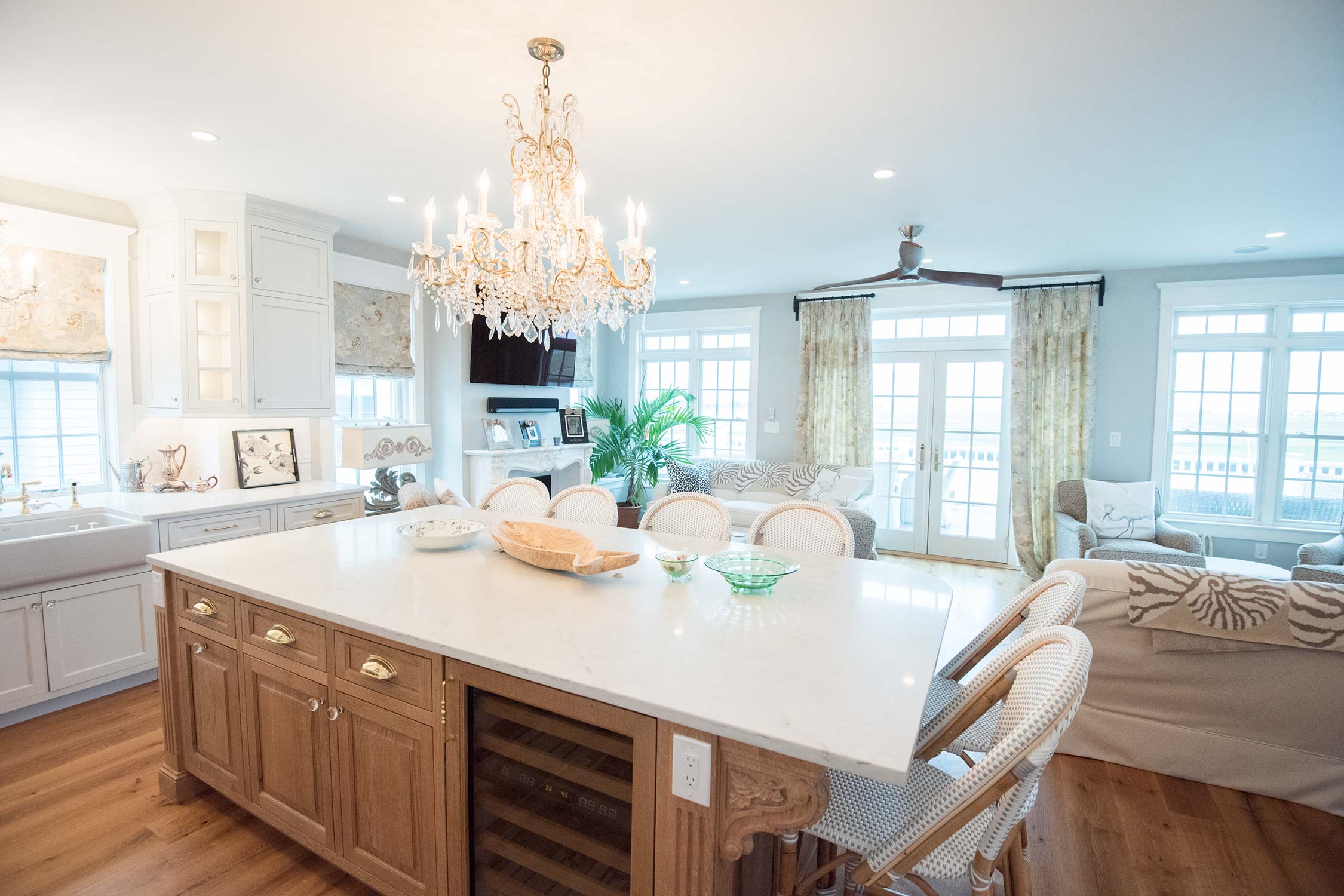 A spacious kitchen with a chandelier, large island, wicker chairs, and an adjacent living area with a sofa, plants, and floral curtains.