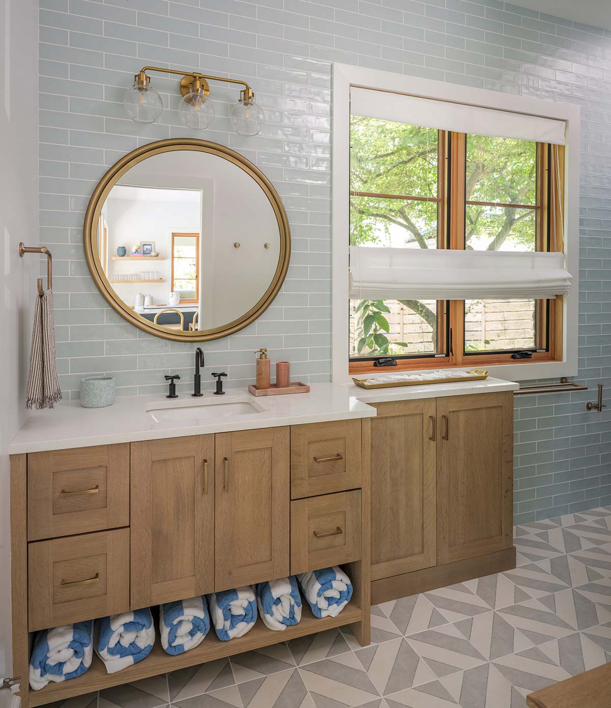 Modern bathroom with light blue tiles, wooden vanity, white sink, round mirror, brass light fixture, and rolled blue towels on a shelf.