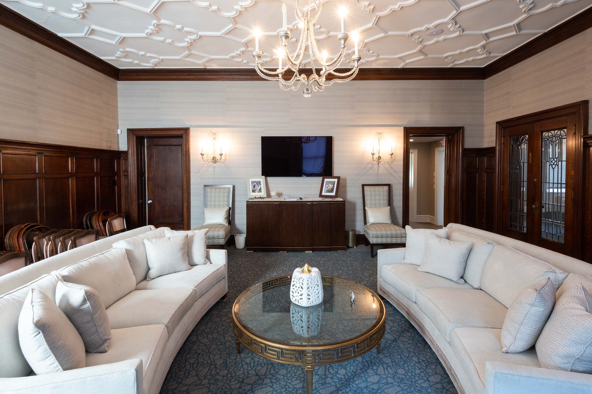 A formal room with a chandelier, wood-paneled walls, and beige sofas. A glass coffee table, TV, and framed photos sit between two armchairs, with a blue patterned rug.