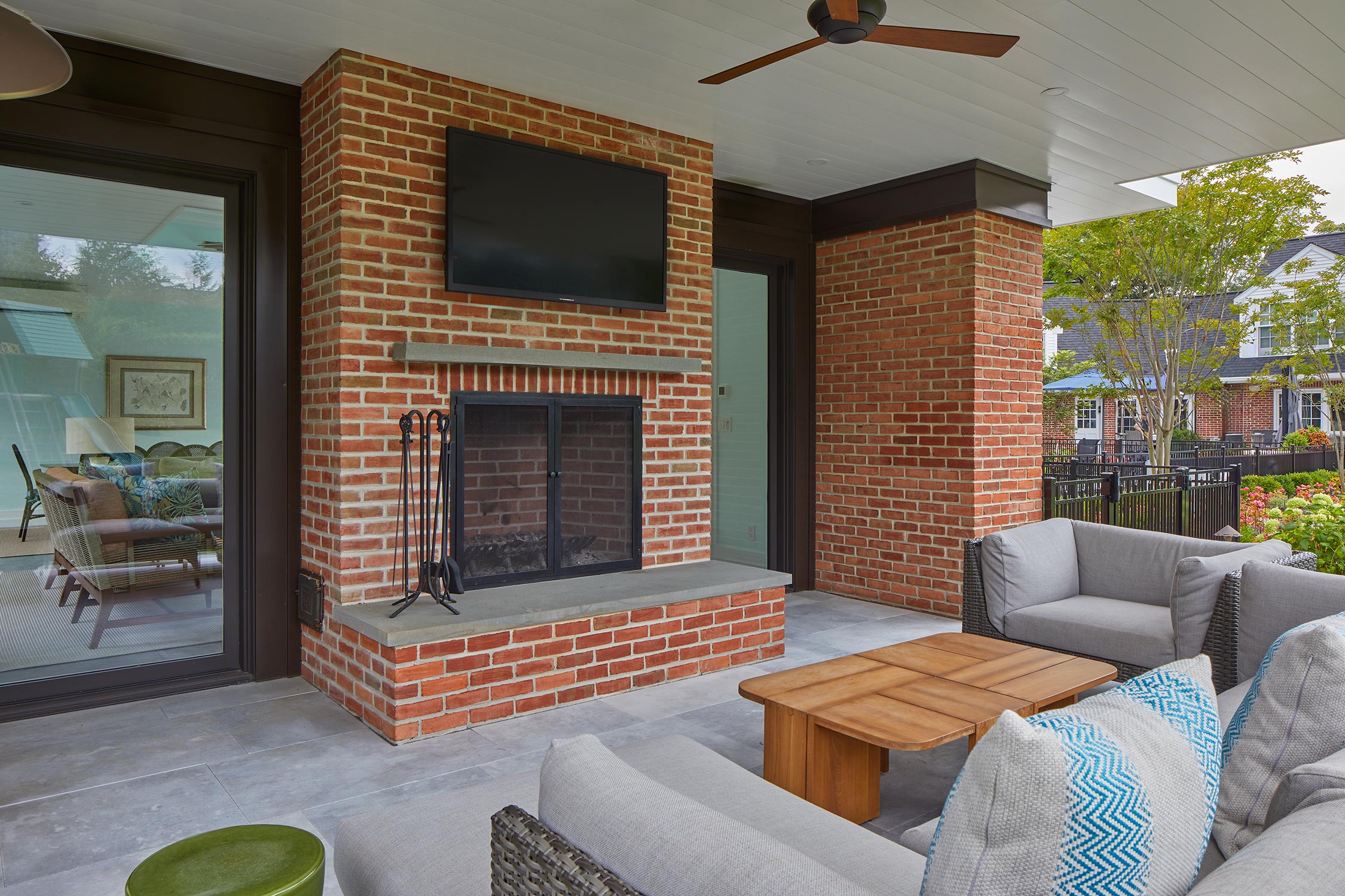 An outdoor patio with a brick fireplace, TV, gray sofas, blue pillows, and a wooden coffee table. Large windows show a cozy interior and greenery outside.