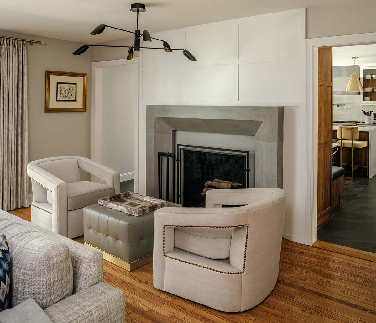Living room with wooden floor, white armchairs, and a gray ottoman. A stone fireplace with a white paneled wall and modern black chandelier above.