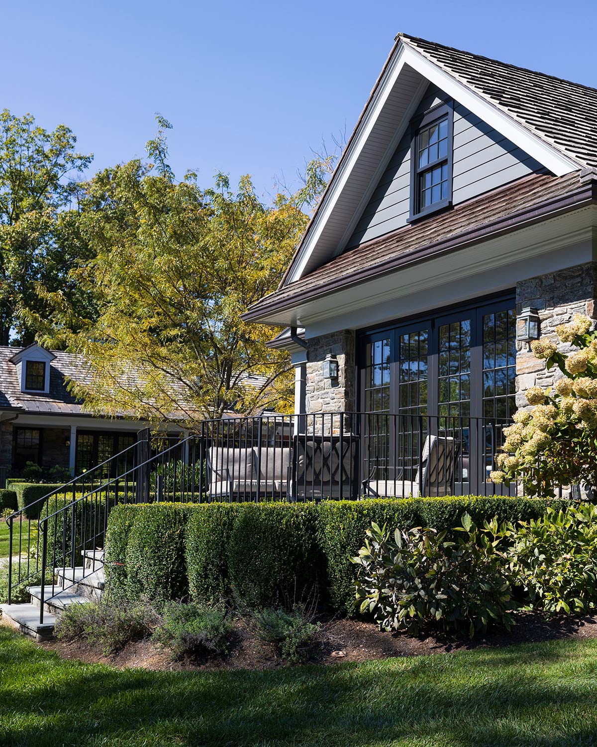 Modern house with gray siding, large glass doors, and a balcony with cushioned chairs, surrounded by lush greenery and a stone staircase.