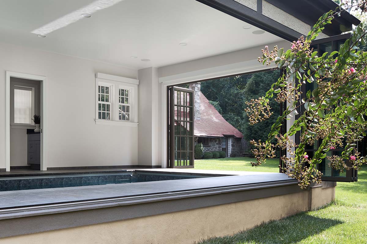 Indoor pool room with large open windows, overlooking a green lawn, blooming shrubs, and a stone house with a red roof.