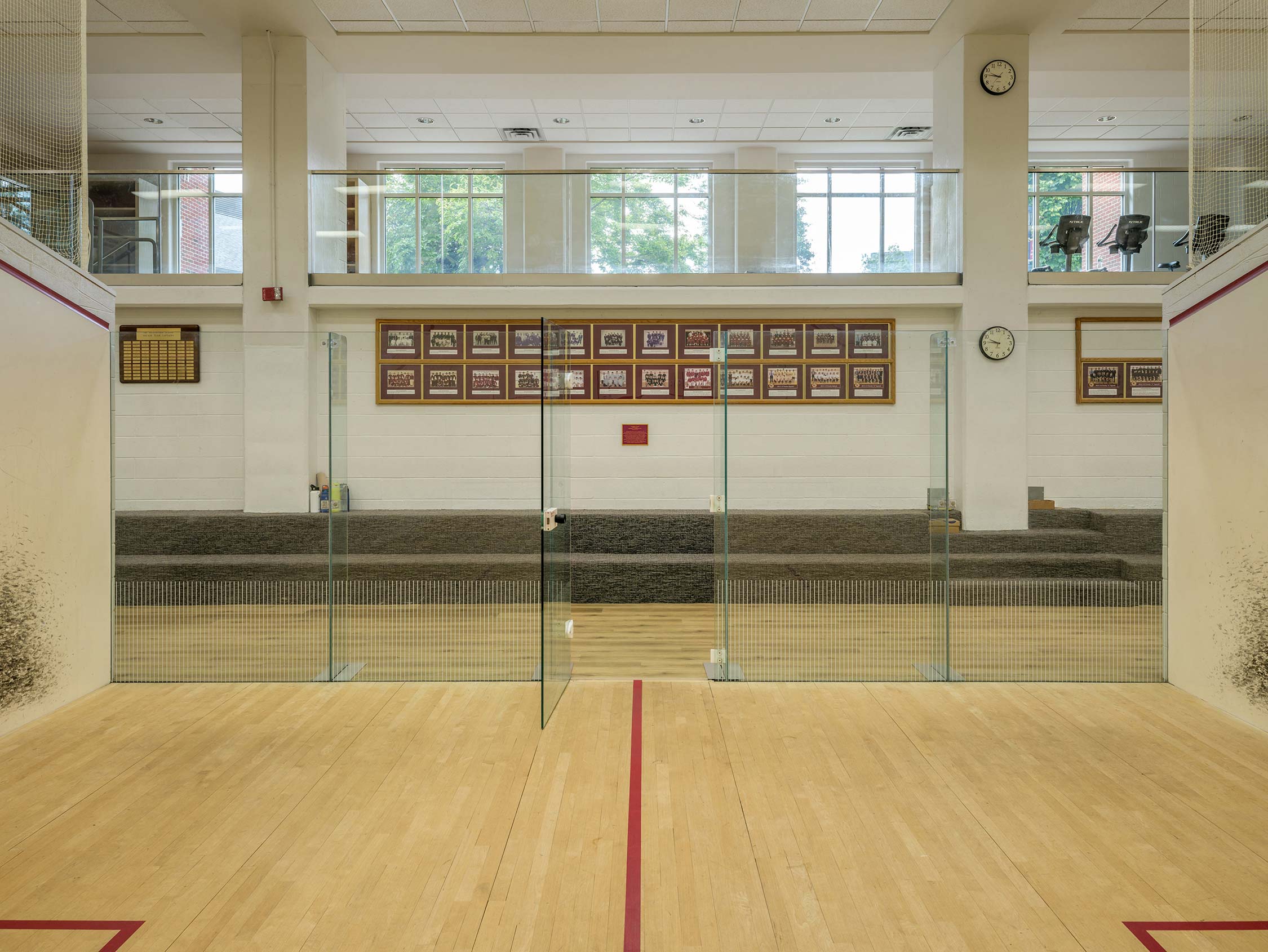 Squash court with glass walls, hardwood floor, and bleachers. Features team photos, awards, and clocks on the wall, with a viewing balcony above.