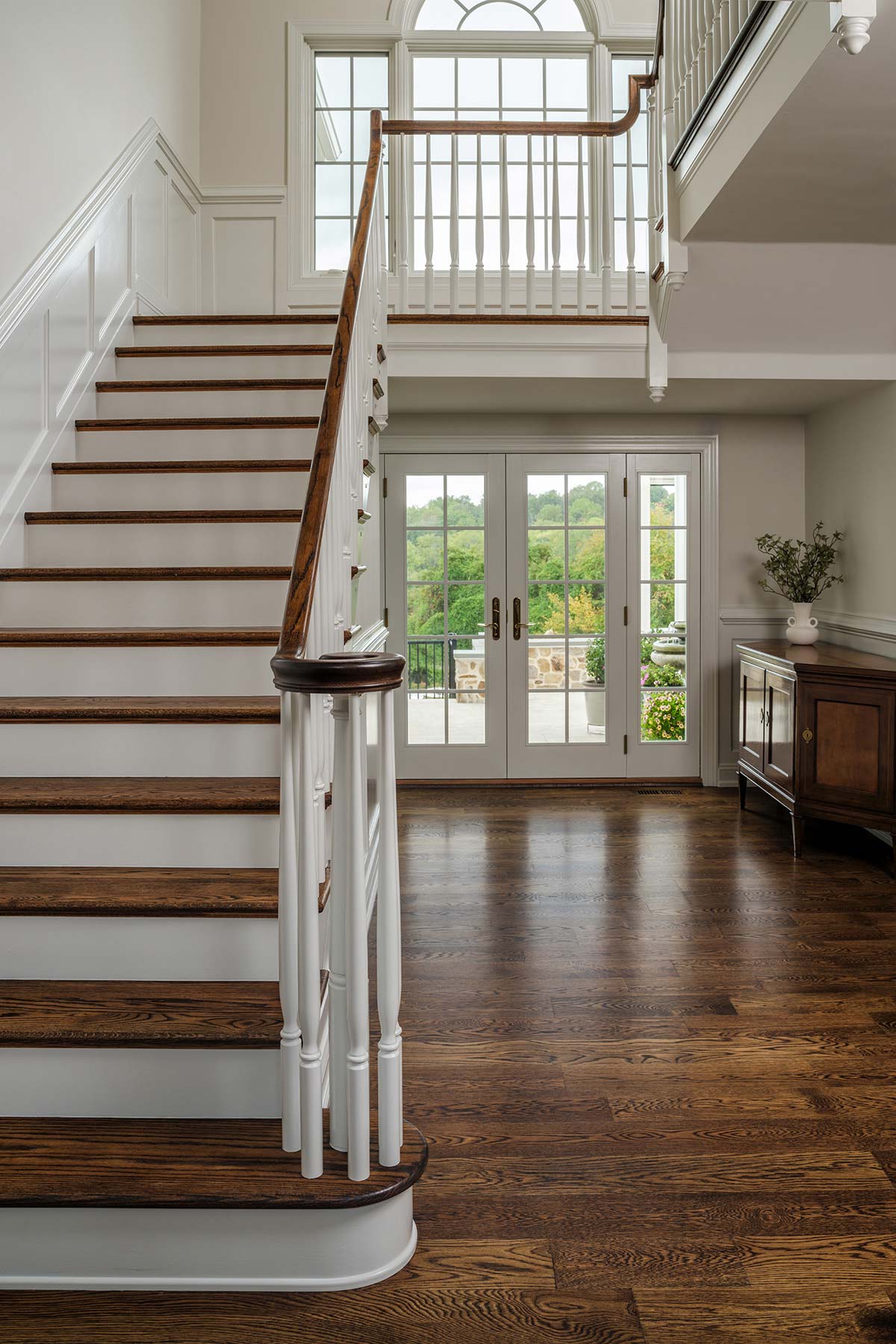 Elegant staircase with wooden steps and white railing, leading to a landing with a large window and glass doors opening to a green view.
