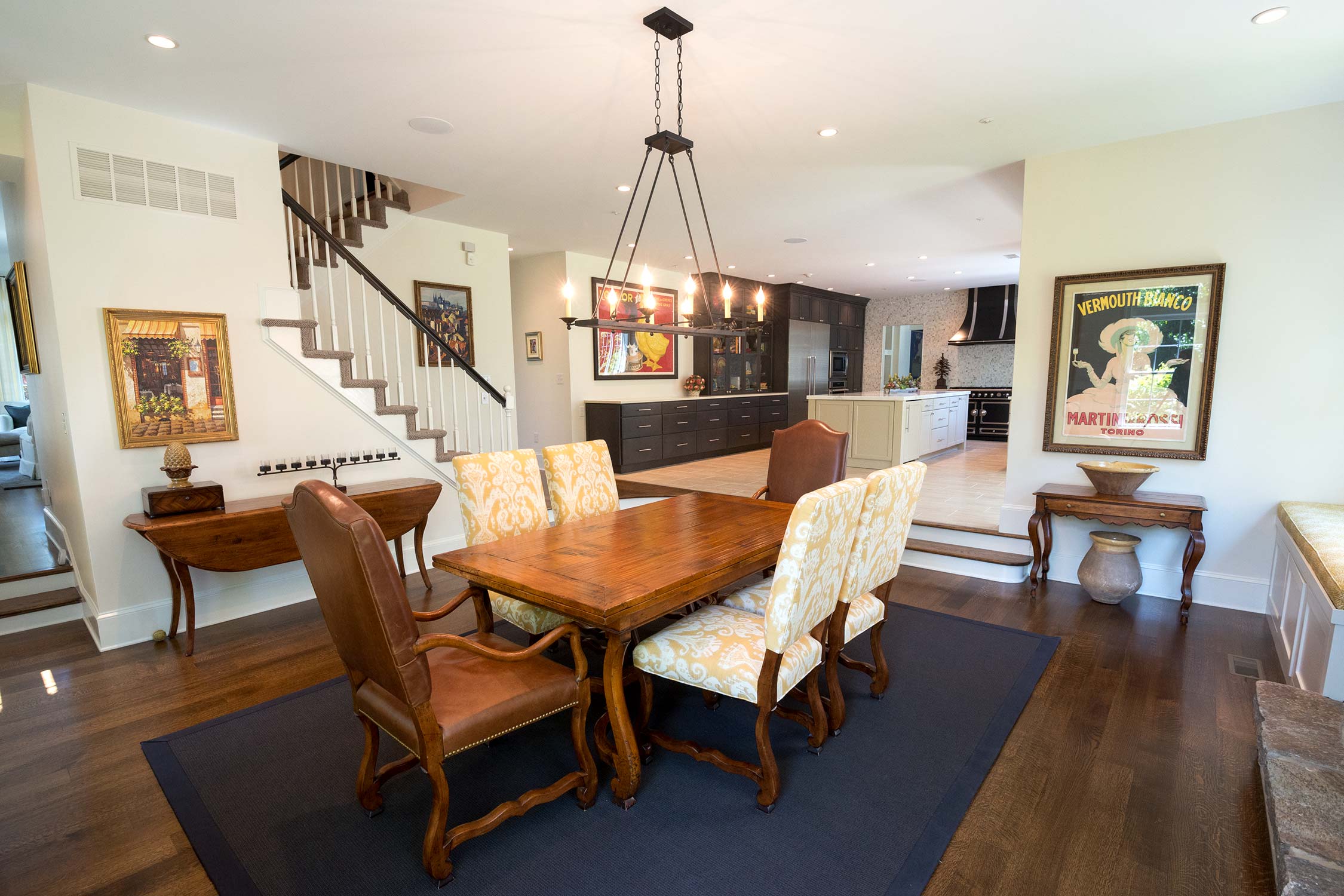 Cozy dining area with wooden table, upholstered chairs, vintage chandelier, and vibrant art, adjacent to a staircase and kitchen.