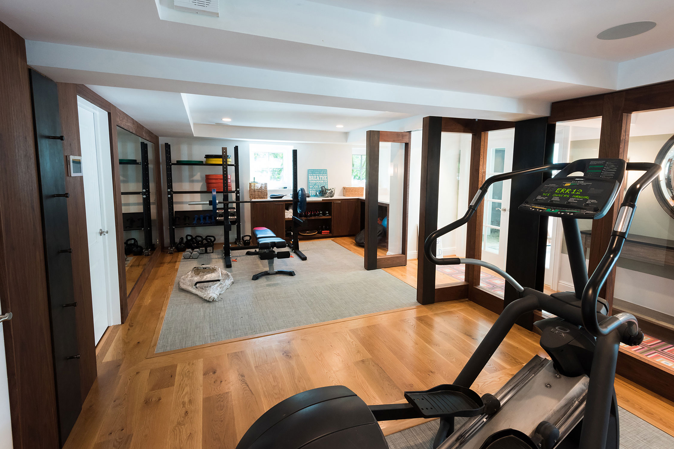 Spacious home gym with a Precor elliptical, weight bench, and storage racks. Features wood flooring, mirrors, and a cozy rug for a stylish workout space.