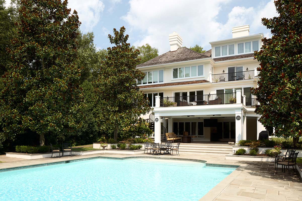 Large white house with pool, patio, and balcony, surrounded by trees and shrubs under a partly cloudy sky.