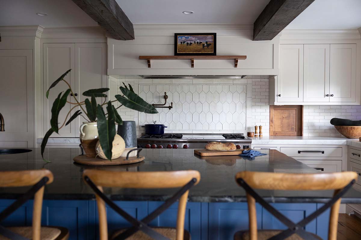 Modern kitchen with blue island, wooden stools, white cabinets, marble backsplash, and exposed wooden beams with a painting.