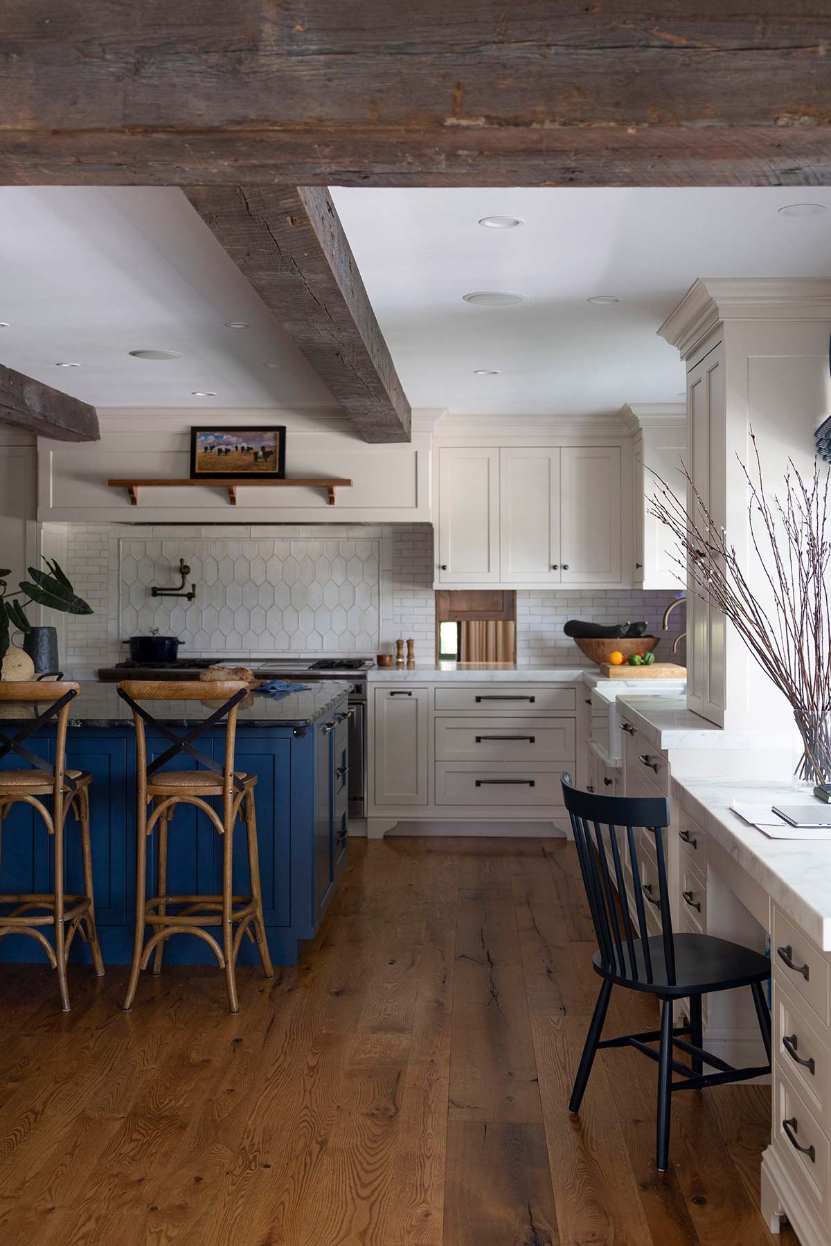 Modern kitchen with a blue island, wooden beams, and white cabinetry. Features a hexagon tile backsplash, rustic wood flooring, and stylish seating.