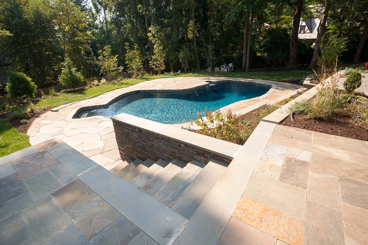 Scenic backyard with a sparkling pool, stone patio, and terraced garden, nestled among lush trees under a bright sky.