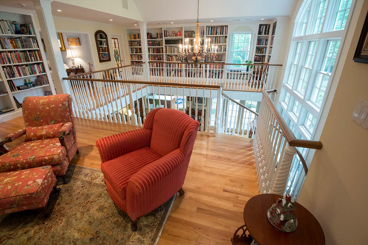 Inviting room with a high ceiling, chandelier, and extensive bookshelves. Features cozy red chairs, large windows, and elegant decor. Perfect for relaxation.