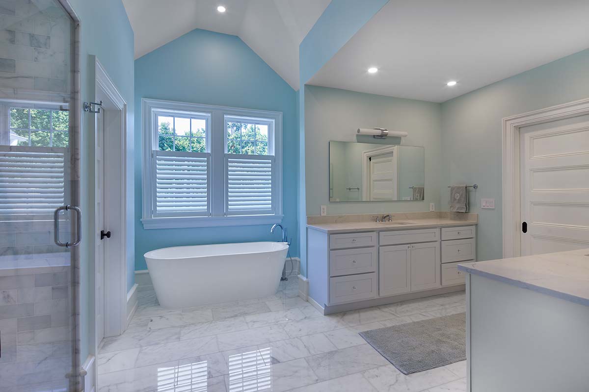 A serene bathroom with light blue walls, a freestanding tub, white vanity, marble floors, a glass shower, and large windows with shutters.