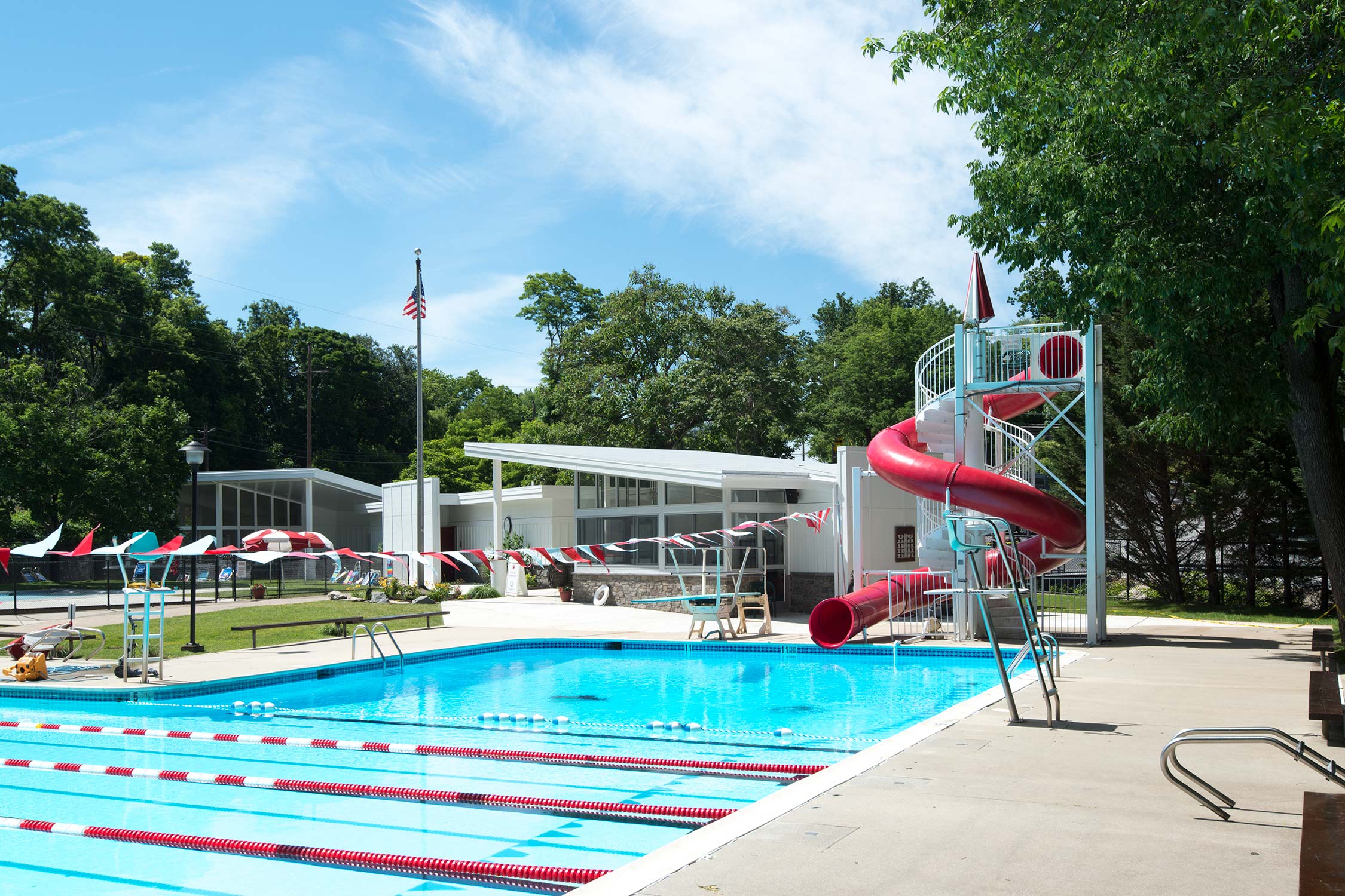 Outdoor pool with red slide, diving board, and lane markers. A lifeguard chair, clubhouse, and flagpole are in the background, surrounded by trees.