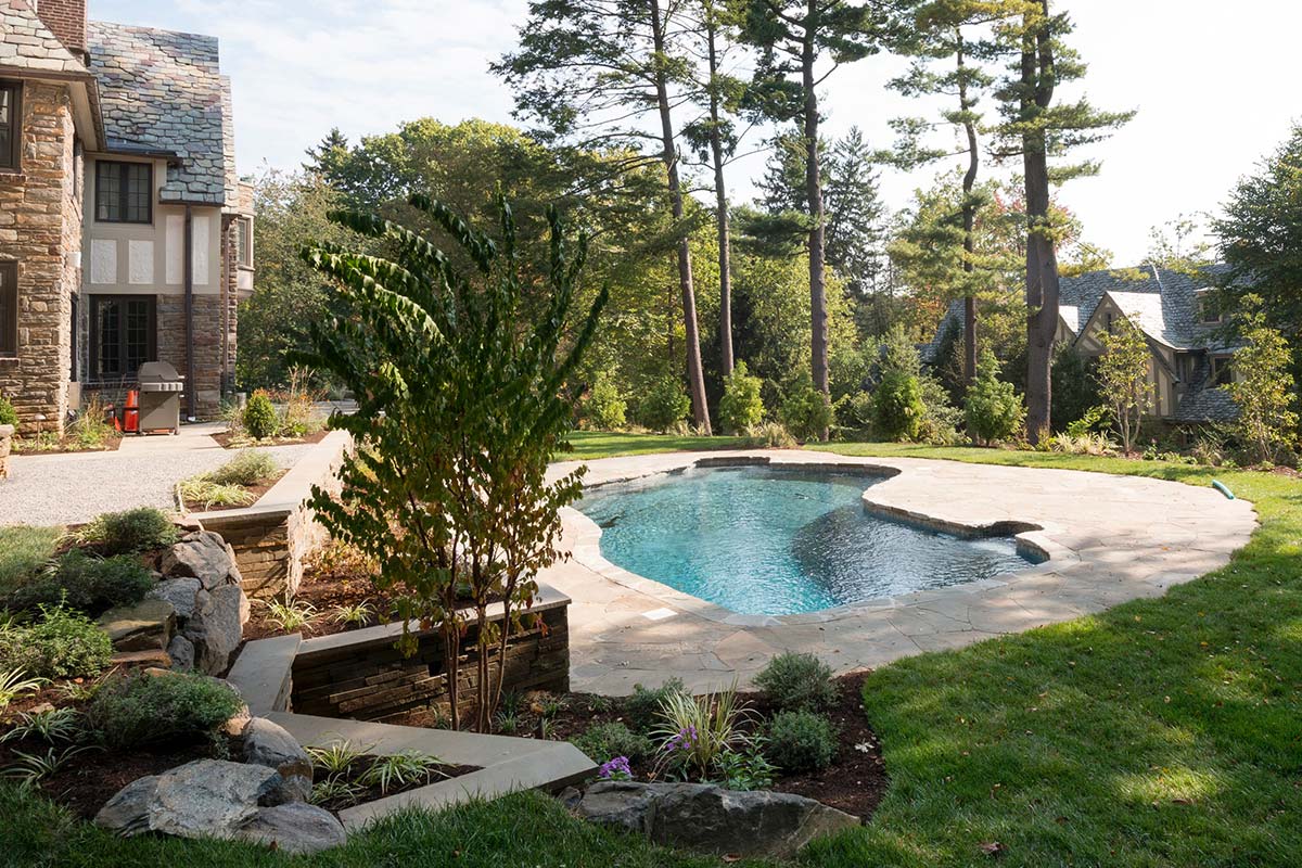 A stone house with a small pool, stone patio, and lush landscaping, surrounded by tall trees and greenery.