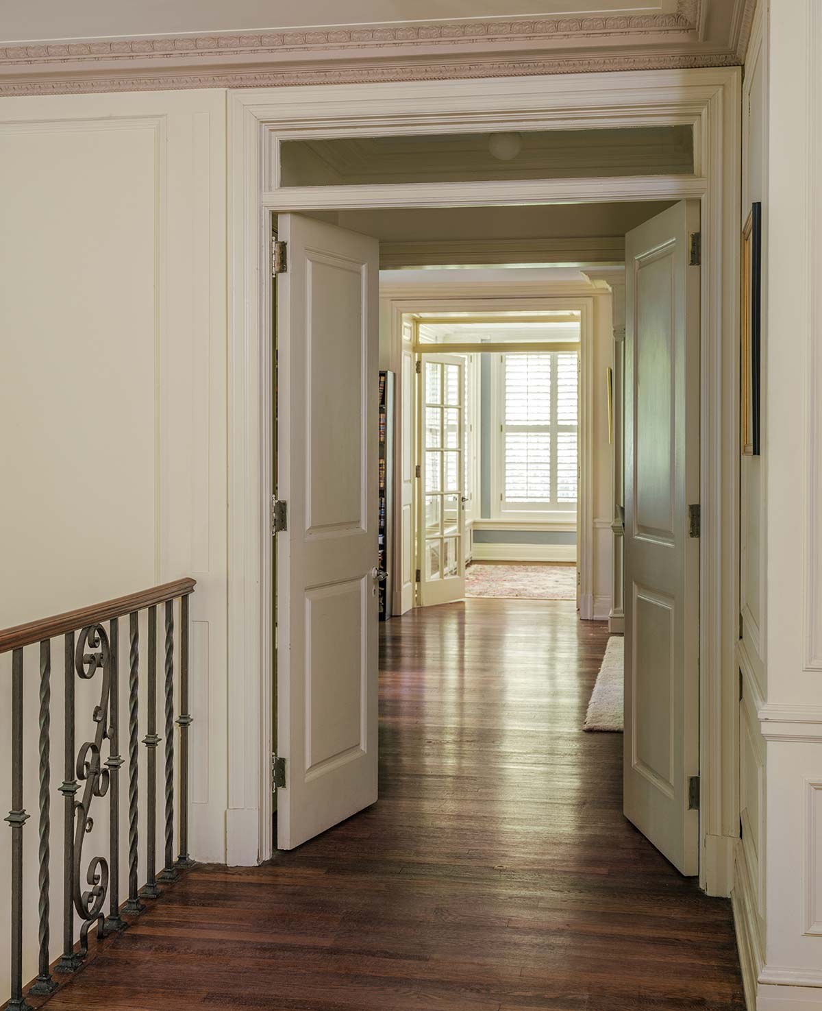 Hallway with open double doors, hardwood floor, and wrought iron railing. Leads to a bright room with large windows and a rug.