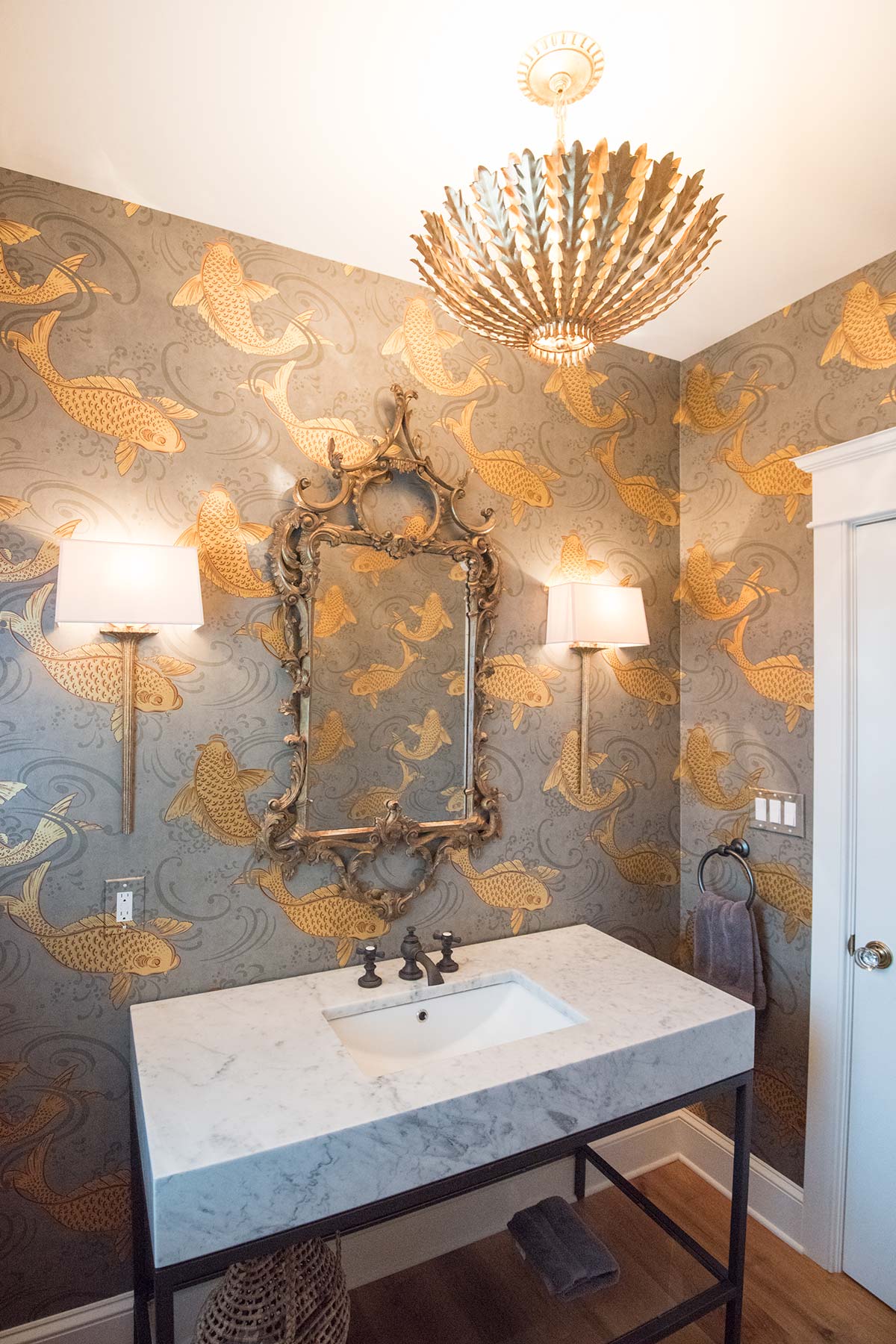 A bathroom with a white marble sink, ornate mirror, gold fish wallpaper, two wall sconces, and a modern gold chandelier.