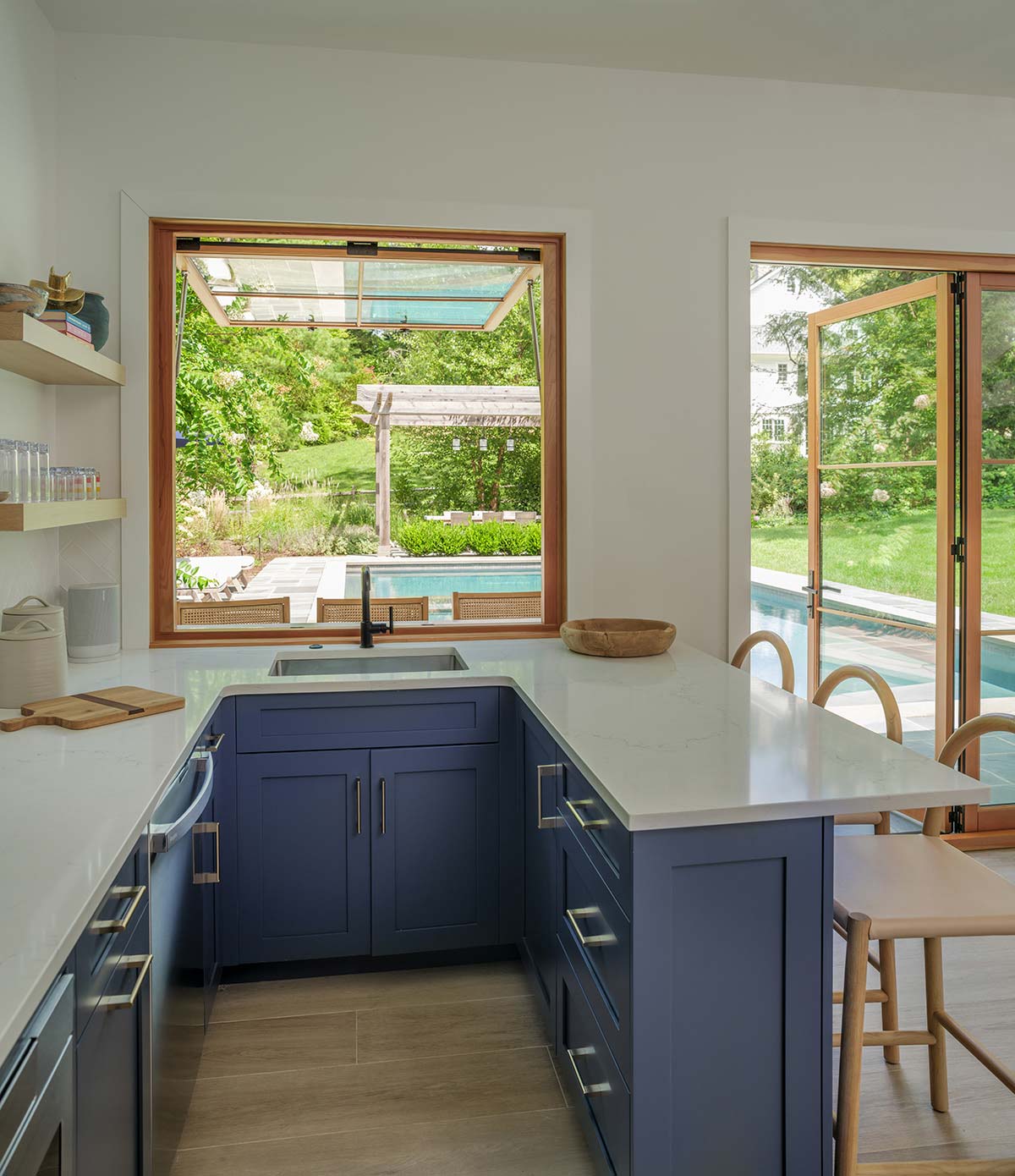 Kitchen with navy cabinets, white counter, sink, and bar stools. Open window and glass doors reveal a pool and lush greenery outside.