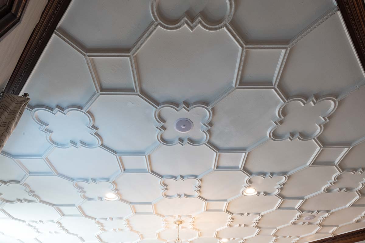 An ornate ceiling with intricate white molding in a geometric pattern, featuring recessed lights and a central circular detail.