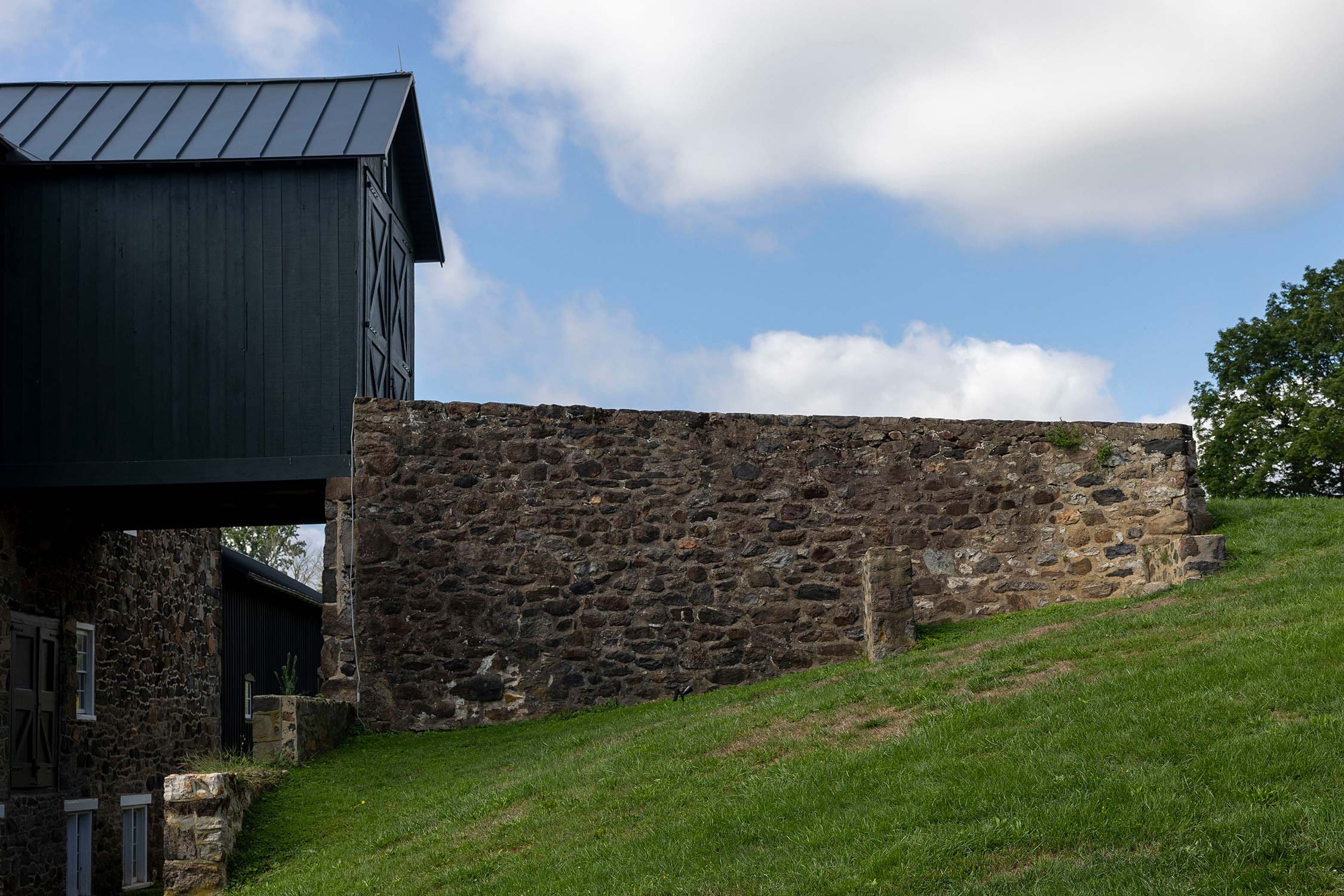 Black barn with metal roof next to a stone wall, on a grassy hill under a partly cloudy sky with trees in the background.