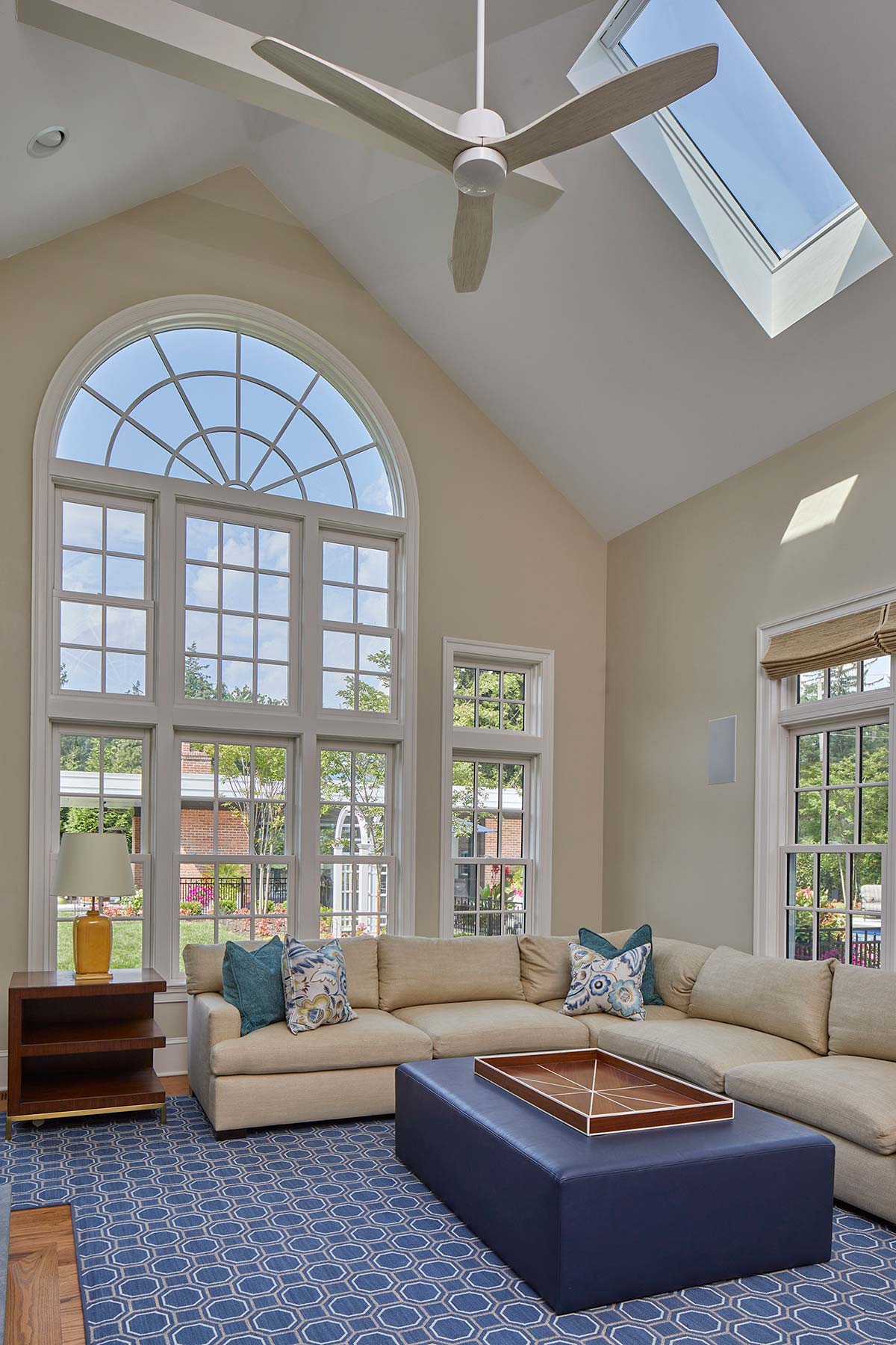 A sunlit living room with a beige sofa, blue pillows, and a patterned rug. Large arched windows and a skylight reveal a green lawn outside.