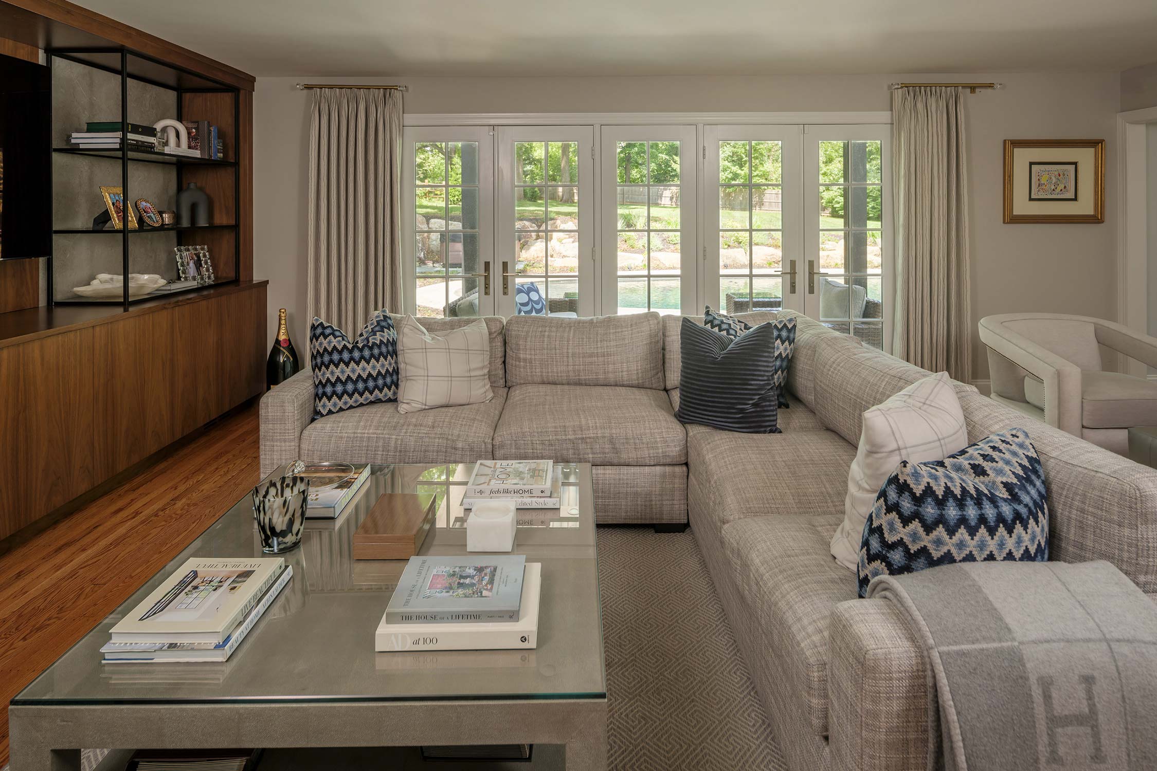 Living room with a gray sectional sofa, blue pillows, and a glass coffee table with books. Large windows show a pool outside, wooden shelves on the left.
