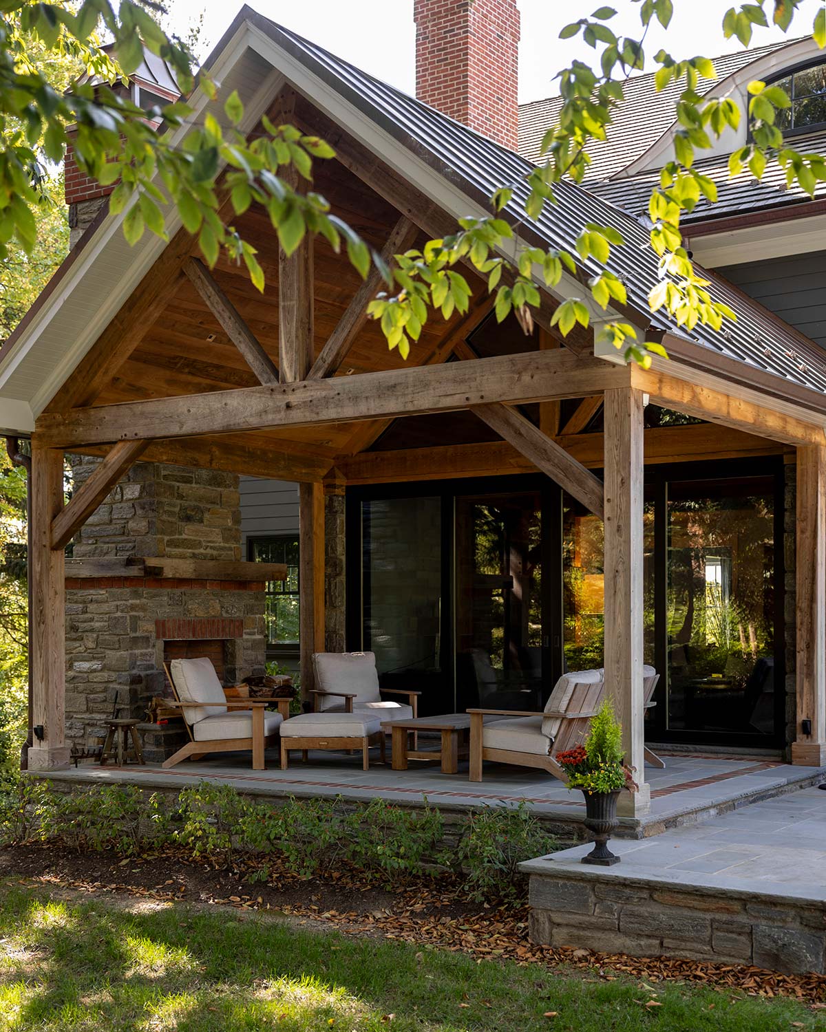 Covered patio with wooden beams, stone fireplace, and cushioned chairs, adjacent to a house with glass doors and lush greenery.