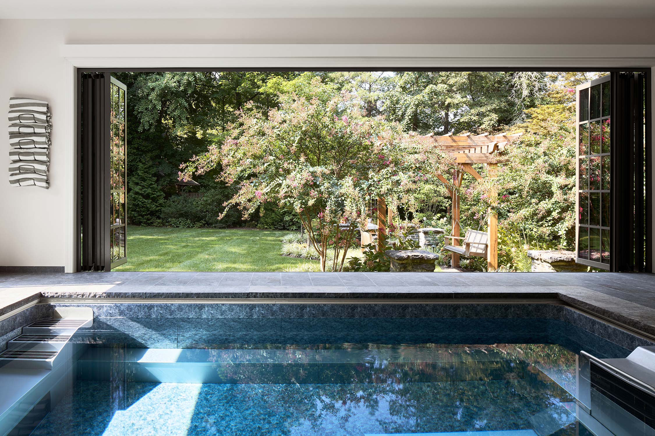 Indoor pool with large open windows, showing a garden with a wooden pergola, swings, green lawn, and blooming shrubs.
