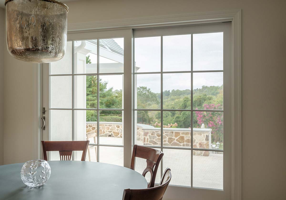Dining area with a round table, wooden chairs, and a glass door offering a view of lush greenery and a stone wall outside.