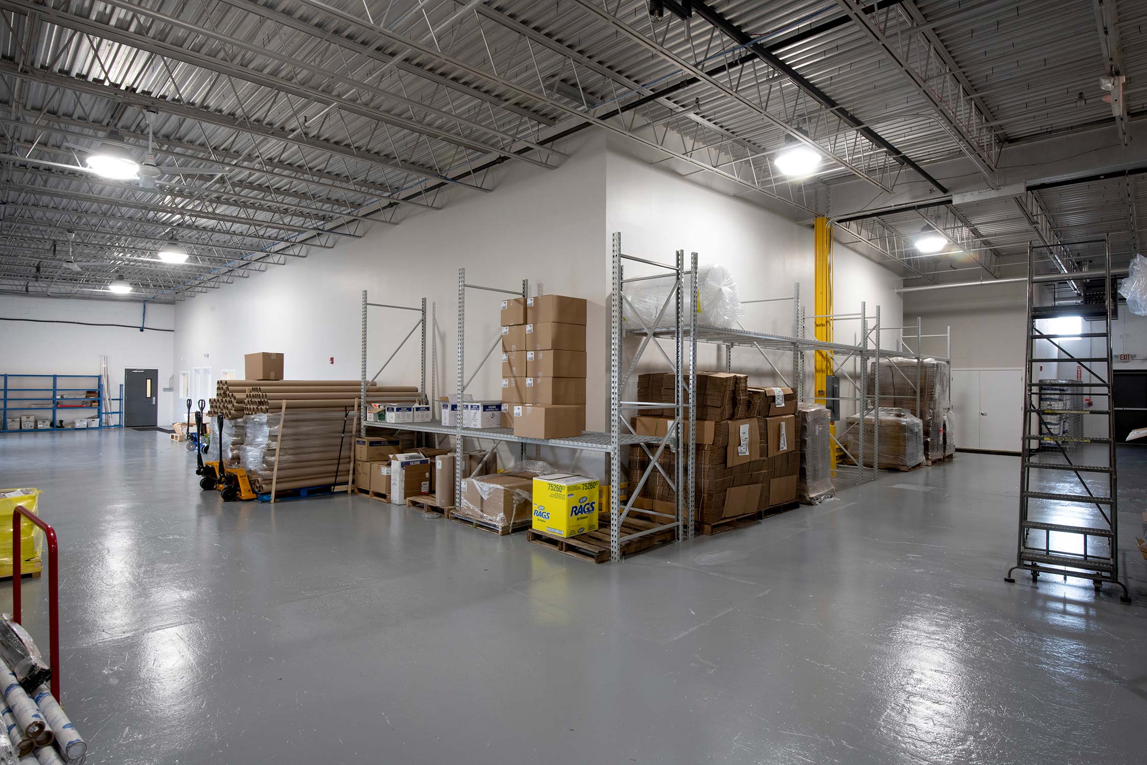 Warehouse with shelves of boxes, pallet truck, and rolls of paper under bright lights.