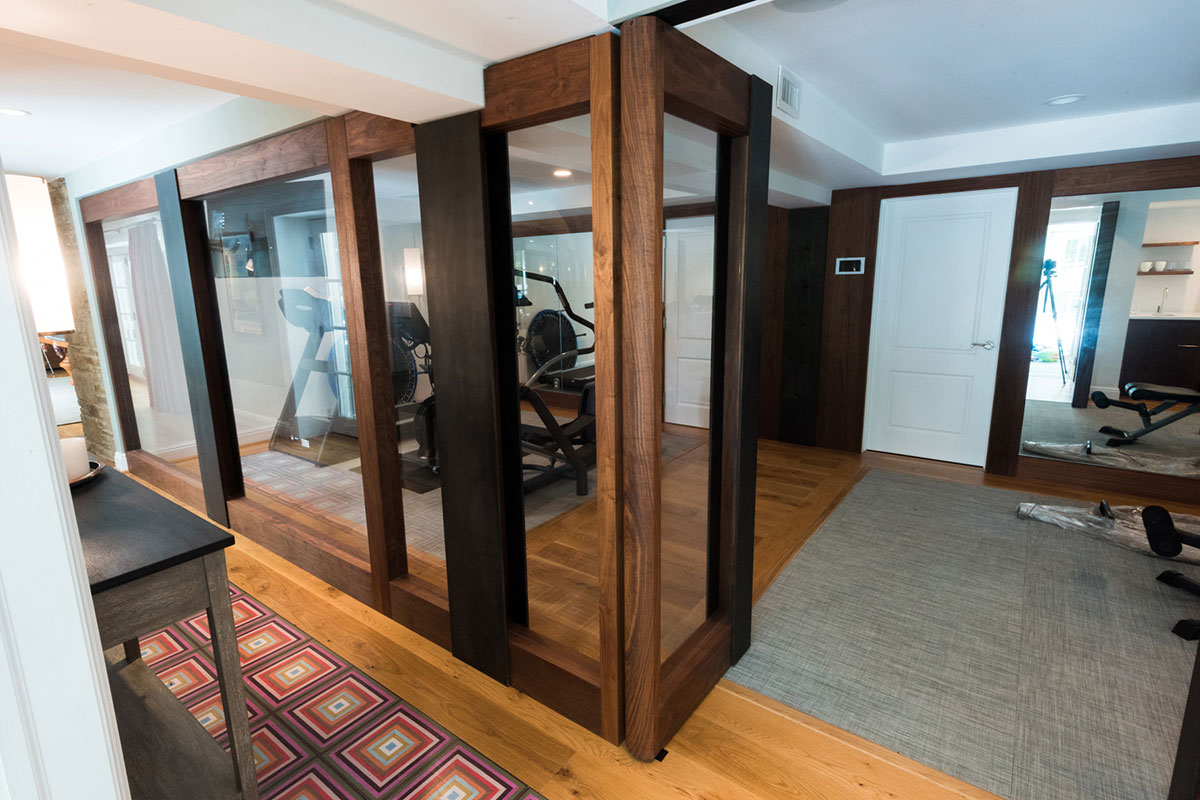 Sleek home gym with a Precor elliptical and weight bench, enclosed by glass walls. Features wood flooring, a colorful rug, and modern design elements.