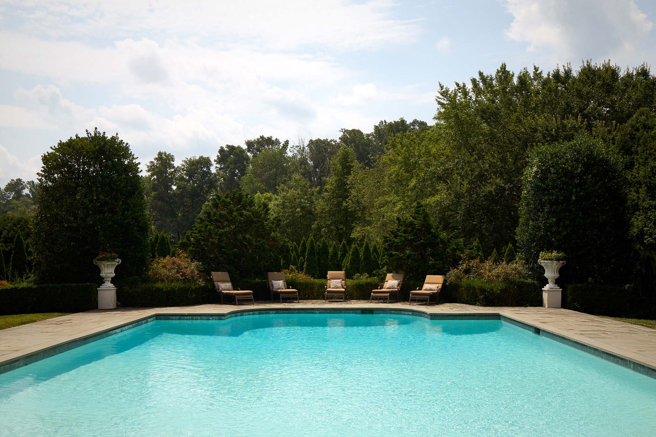 A backyard with a rectangular pool, stone patio, lounge chairs, decorative urns, and dense greenery.