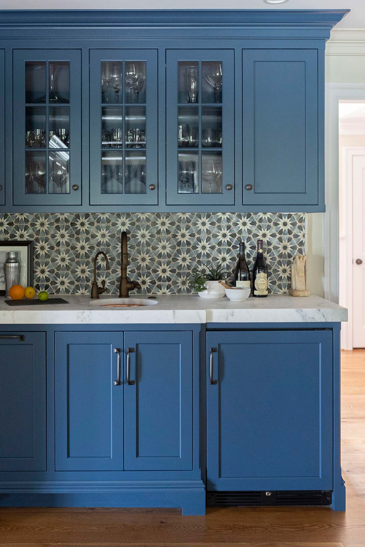 Blue kitchen cabinets with glass doors, star-patterned tile backsplash, and marble countertop. Includes sink, wine bottles, and fruit.