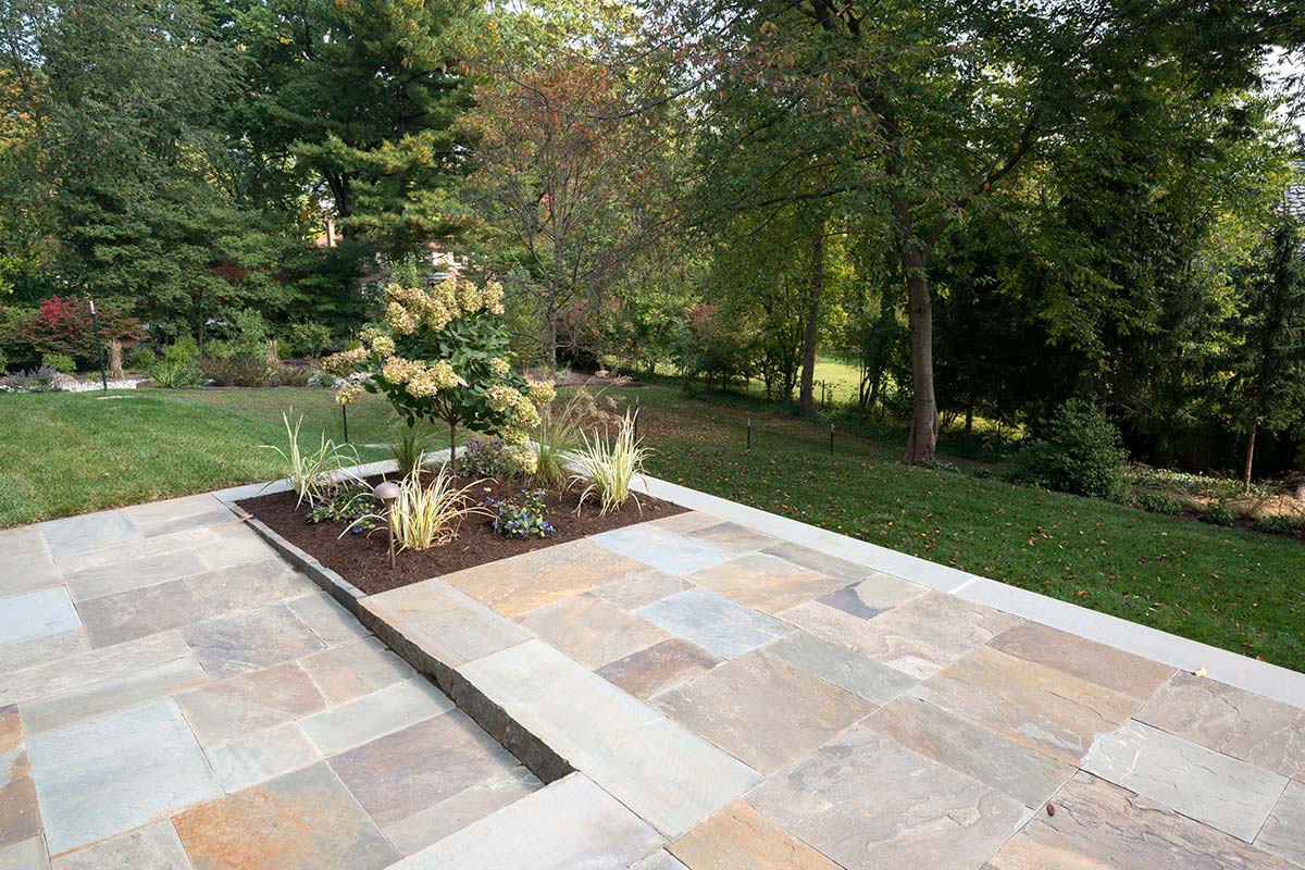Serene patio with stone tiles and a landscaped garden featuring hydrangeas and grasses, surrounded by lush trees.