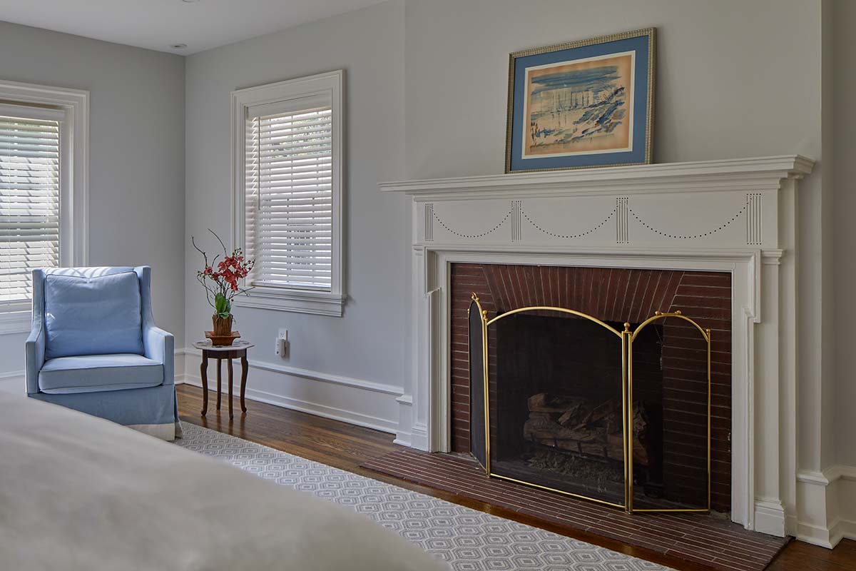 Cozy room corner with a brick fireplace, white mantel, blue armchair, small table with flowers, and large windows with shutters.