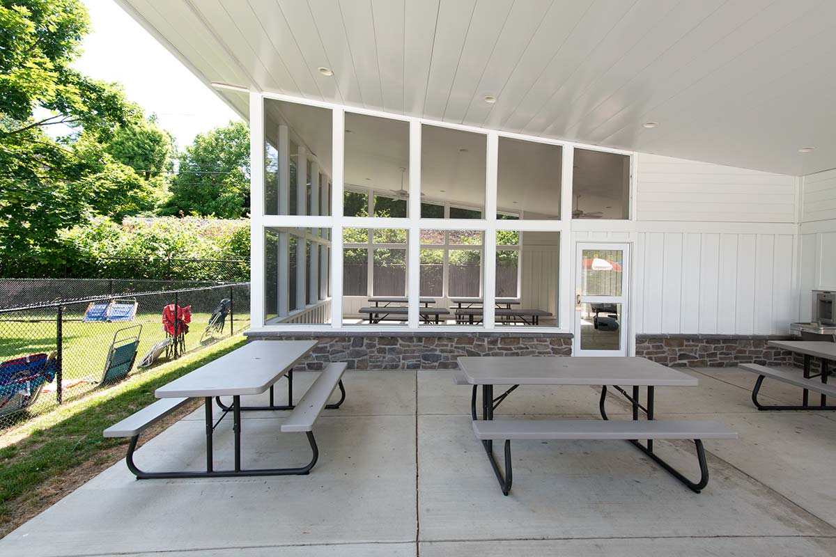 Outdoor patio with picnic tables under a covered roof, next to an office area at a swim club with large windows and a stone wall. A fenced yard with chairs is visible in the background.