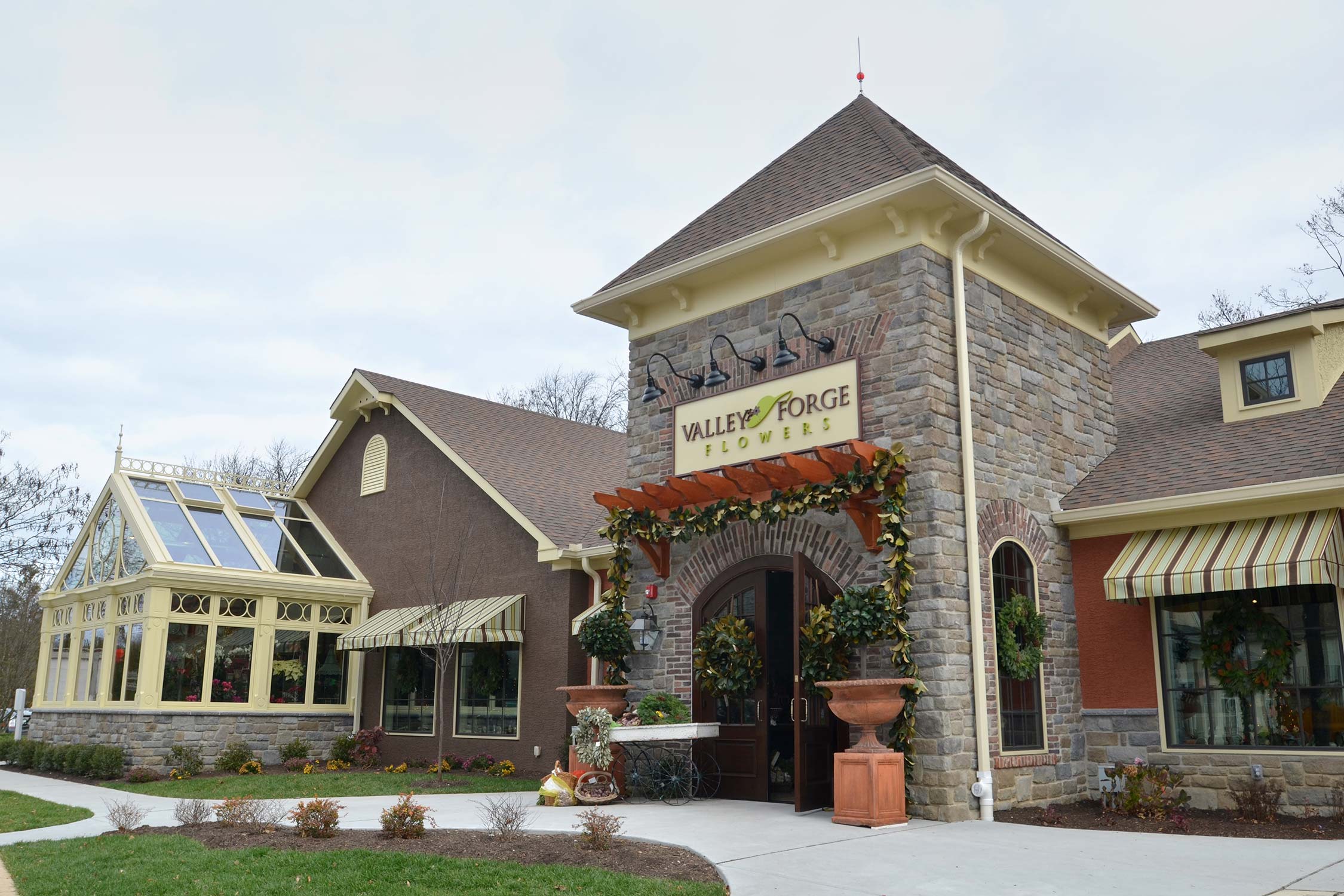 Valley Forge Flowers entrance with stone facade, pergola, wreaths, potted plants, and a wooden cart on an overcast day.