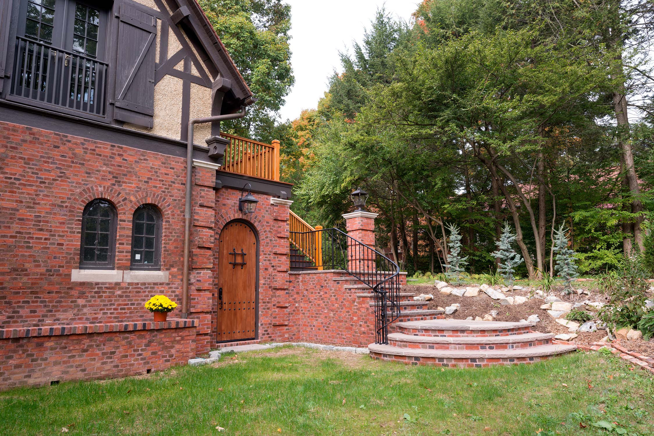 A brick house with a wooden door, arched windows, and a balcony, featuring brick steps and a lawn with trees.