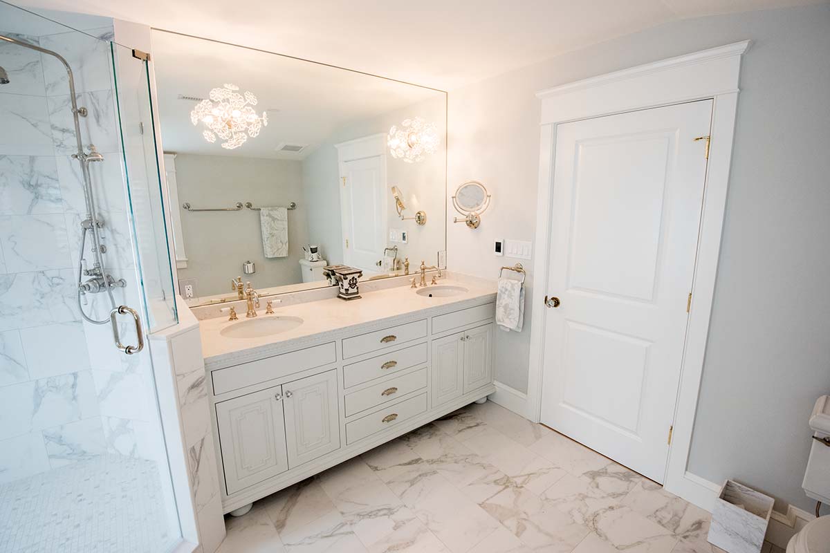A modern bathroom with a double sink vanity, large mirror, chandelier, marble tiles, and a glass shower.