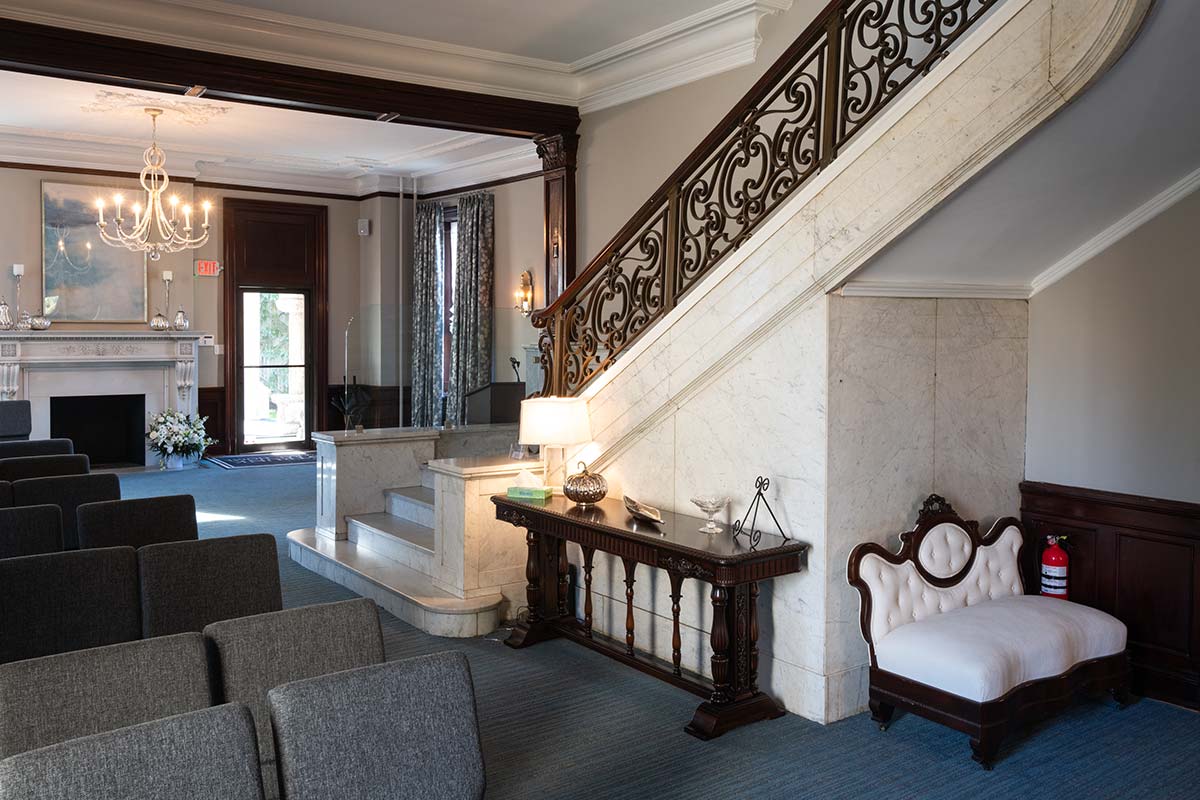 A formal room with a marble staircase, ornate railing, and chandelier. Gray chairs, a white sofa, and a fireplace with flowers are visible, along with blue curtains.