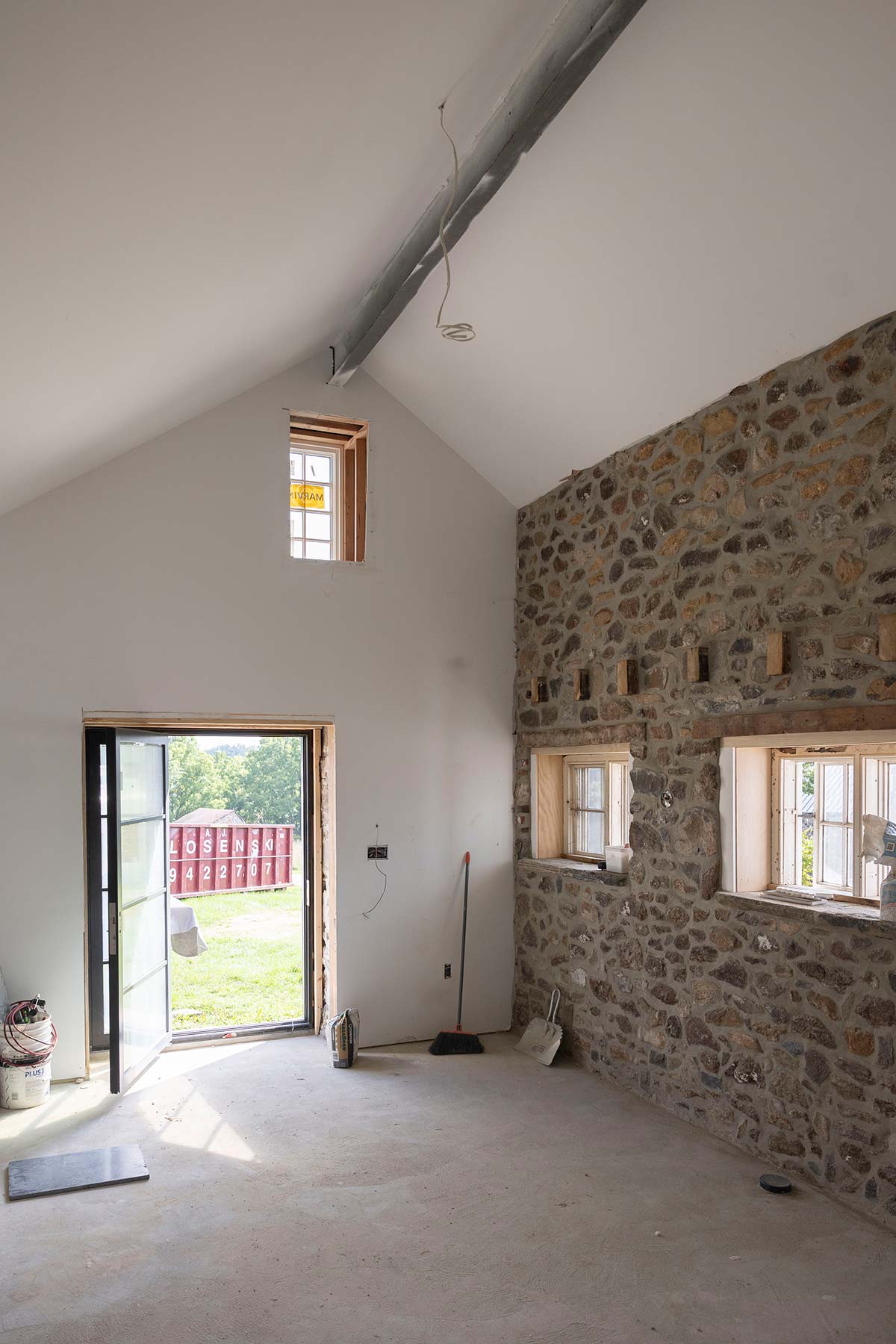 Unfinished room with stone walls, open door, and windows, showing construction tools and a dumpster outside.