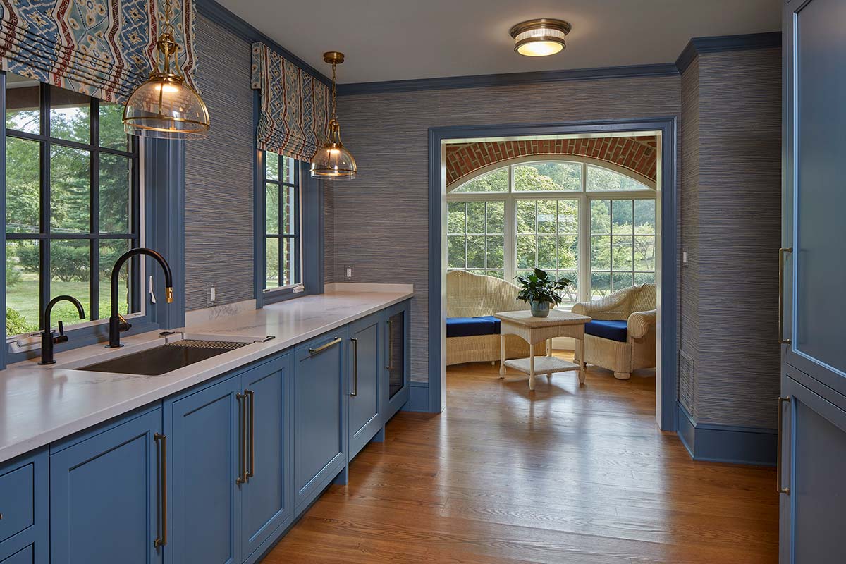 A kitchen with blue cabinets, a sink, and pendant lights. Patterned curtains frame windows, and a cozy sitting area with wicker chairs is visible through a brick archway.