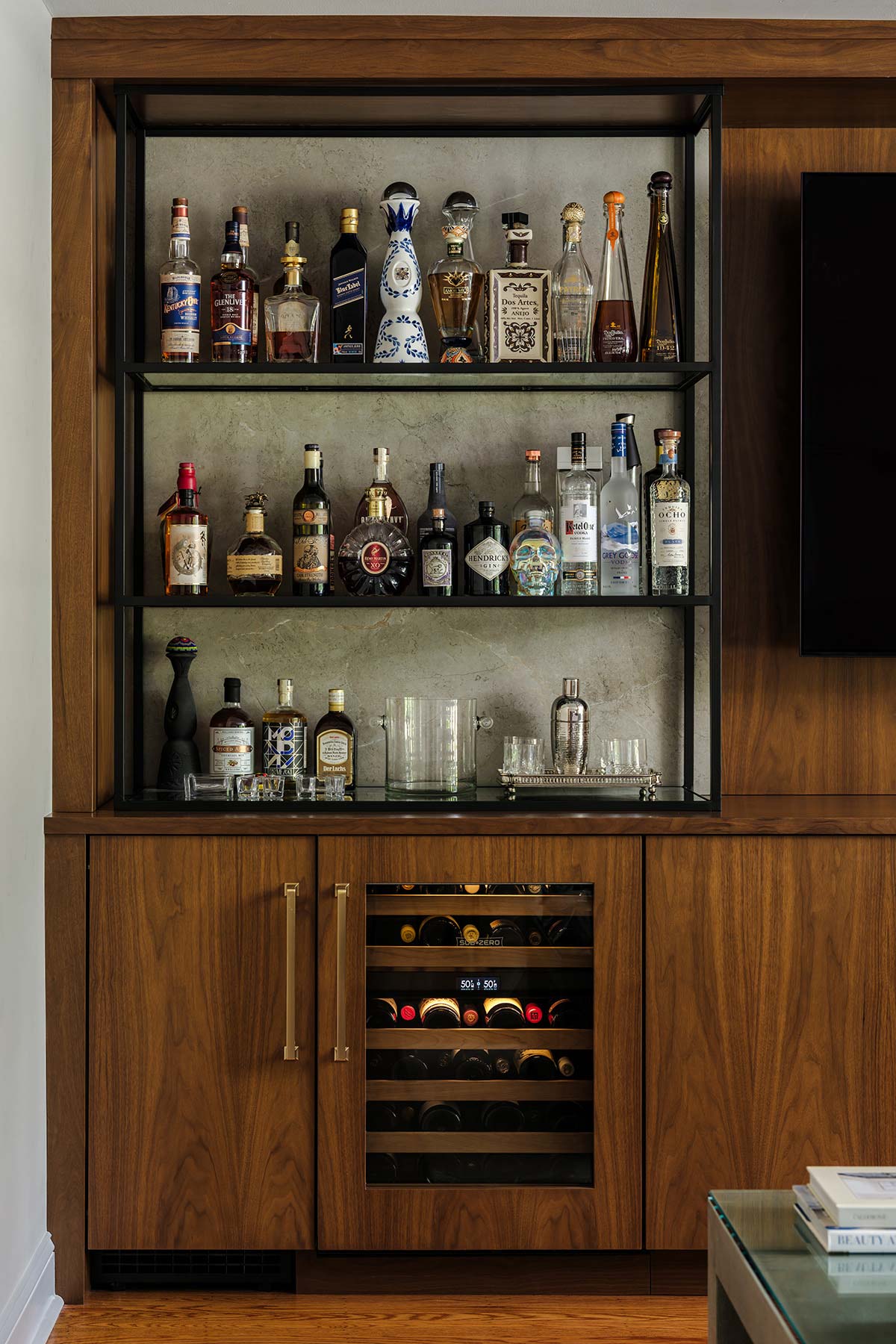 Wooden bar cabinet with open shelves, various liquor bottles, glassware, and a wine cooler. A TV is mounted on the right side.