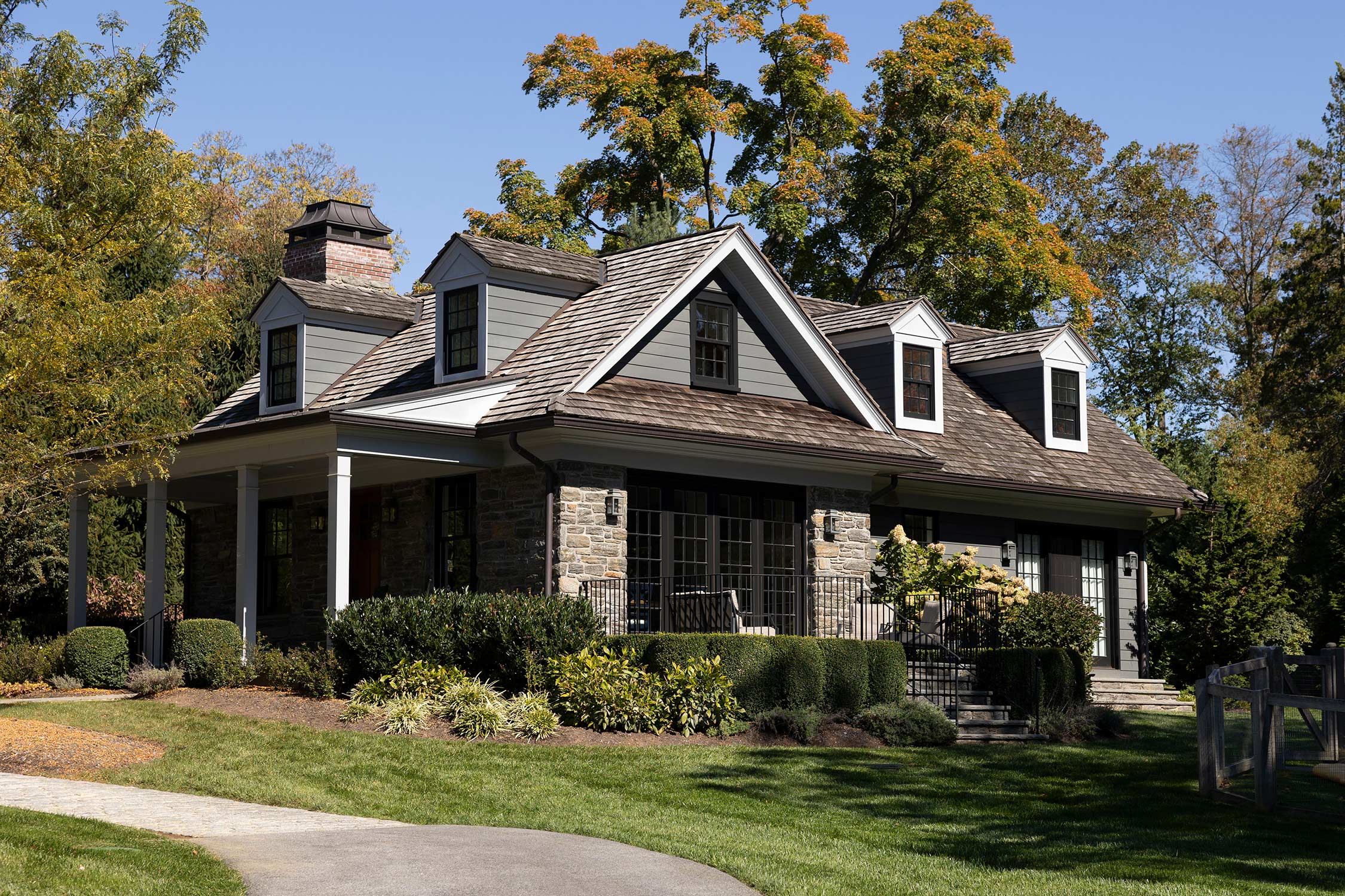 Cozy house with gray siding, shingled roof, and a porch with columns, surrounded by lush greenery, shrubs, and a stone path.