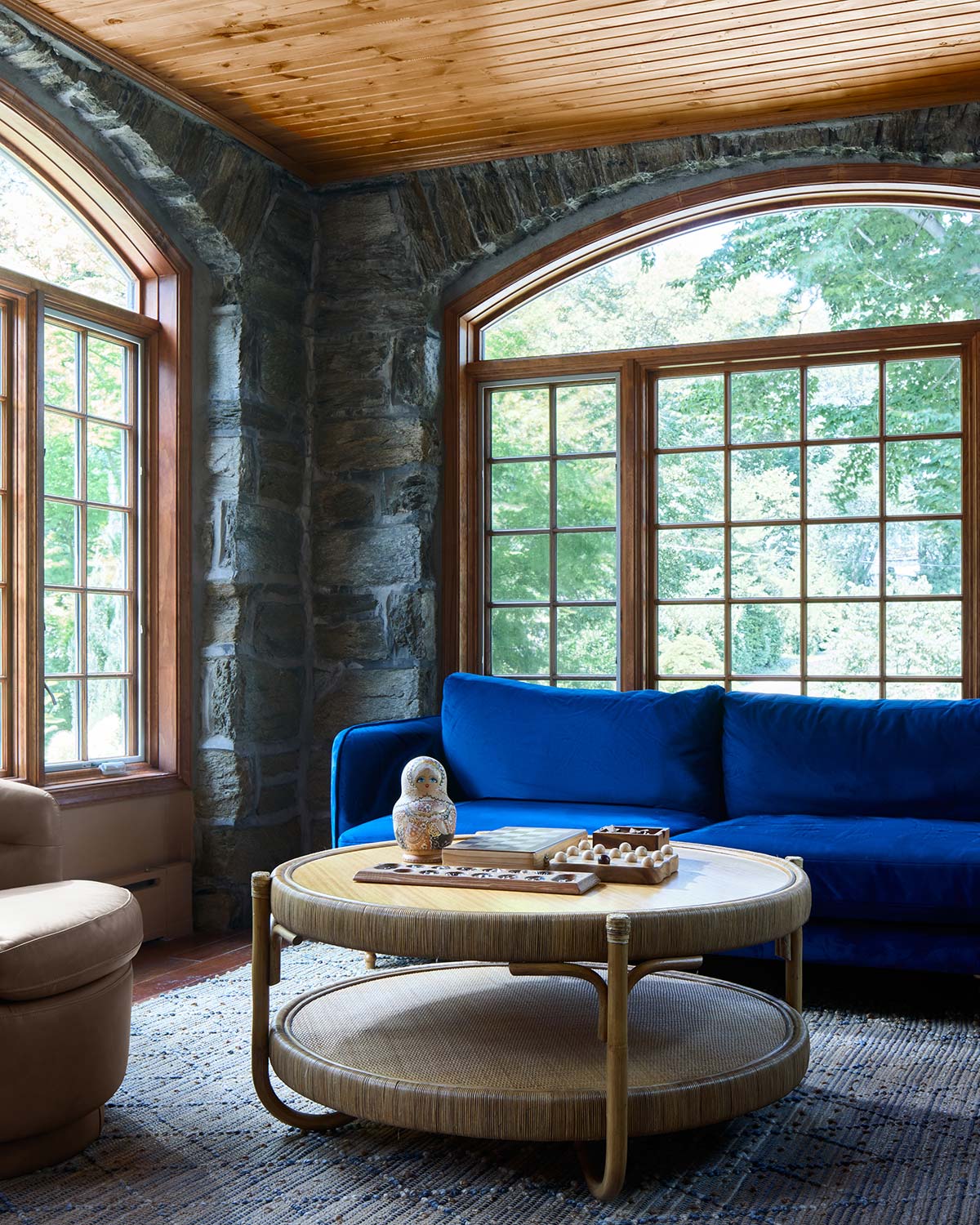 Cozy room with stone walls, wooden ceiling, large windows, blue sofa, round wicker table, and board game.