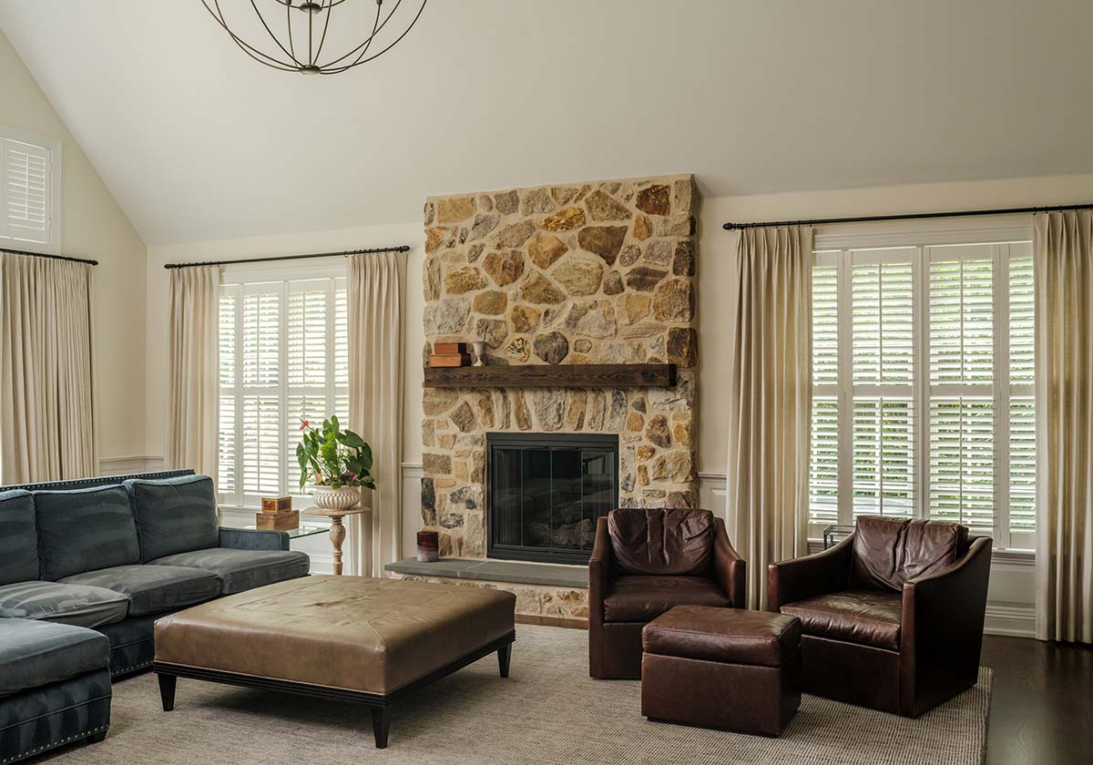 Cozy living room with a stone fireplace, leather chair and ottoman, blue sofa, ottoman, and large windows with shutters.